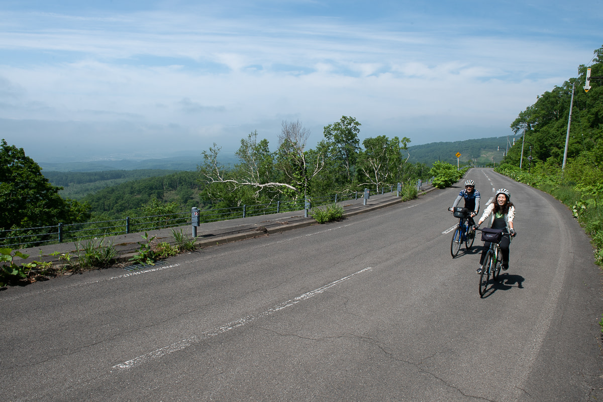 Two cyclists climb up a mountain road with blue skies and white clouds above.