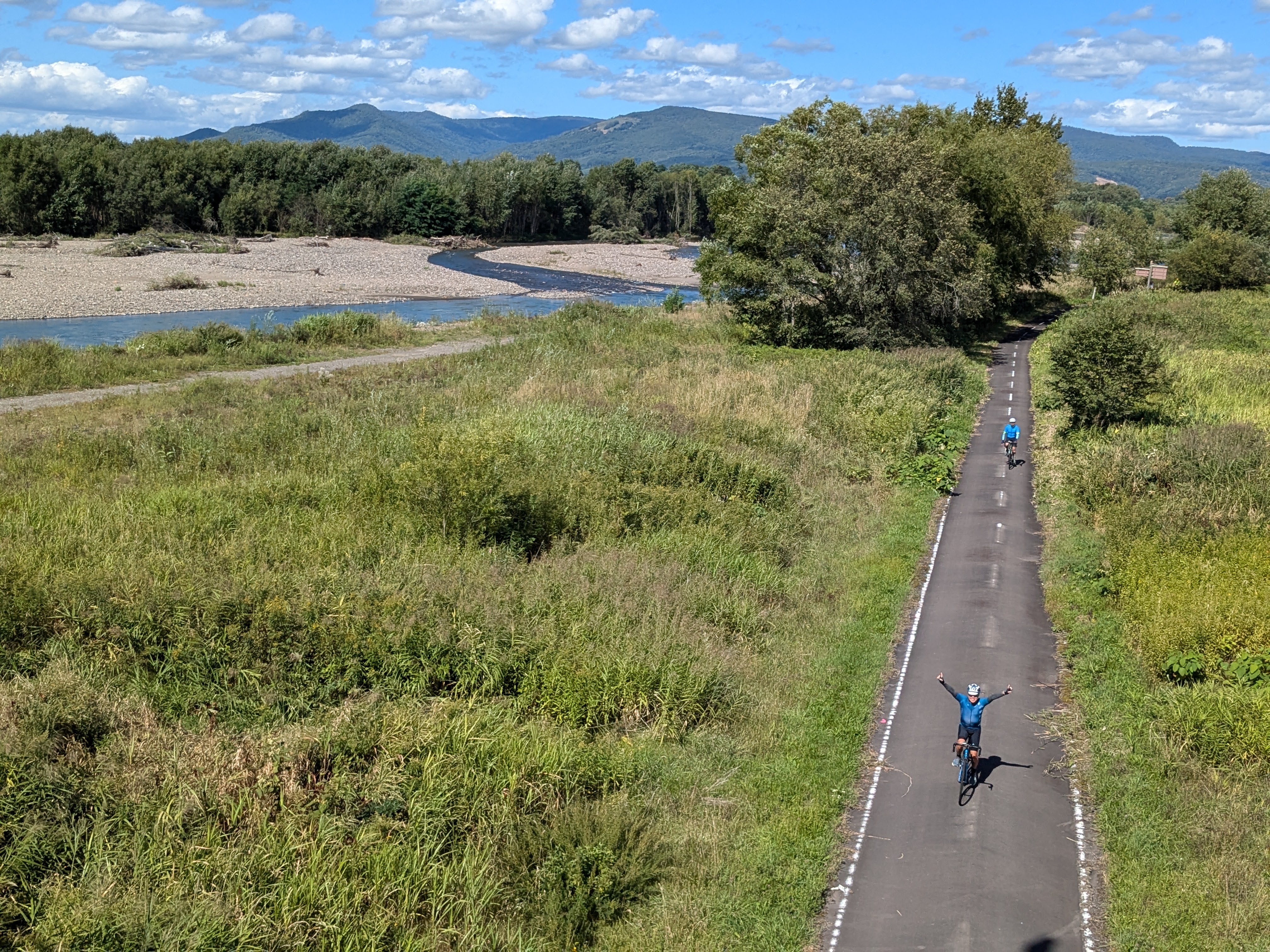 An aerial view of cyclists proceeding down a cycling road. There is a winding river alongside them. It is a beautifully sunny day.