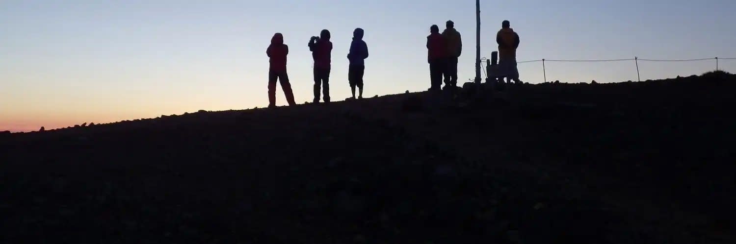 A group of hikers are silhouetted on a mountain top at sunset. The hikers and ridgeline are pitch black, while the ski has the pastel colours of last light.