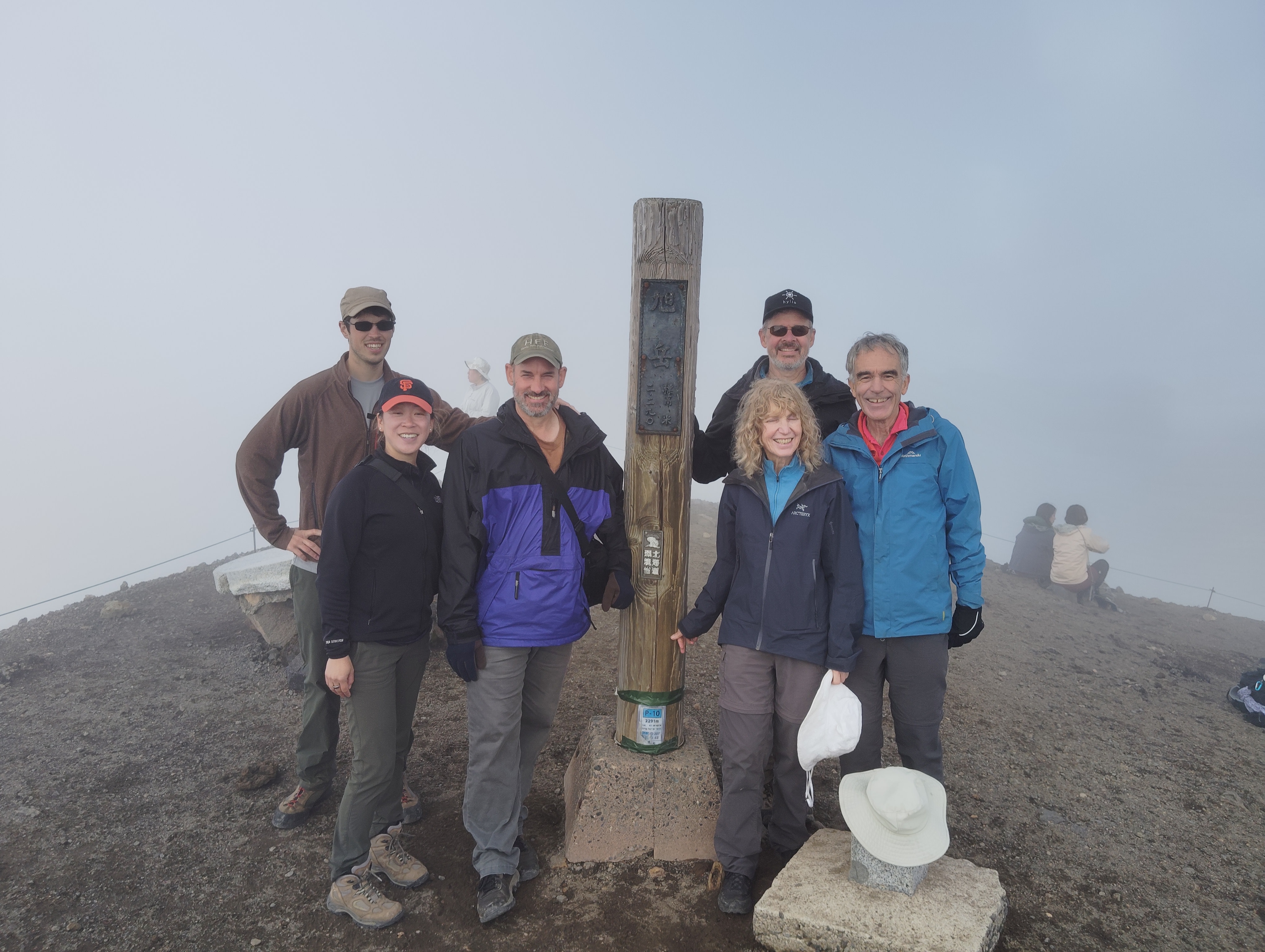 Six hikers smile at the camera at the summit of Mt. Asahidake, Hokkaido. They stand around a pole which reads "Mt. Asahidake, 2290m" in Japanese.