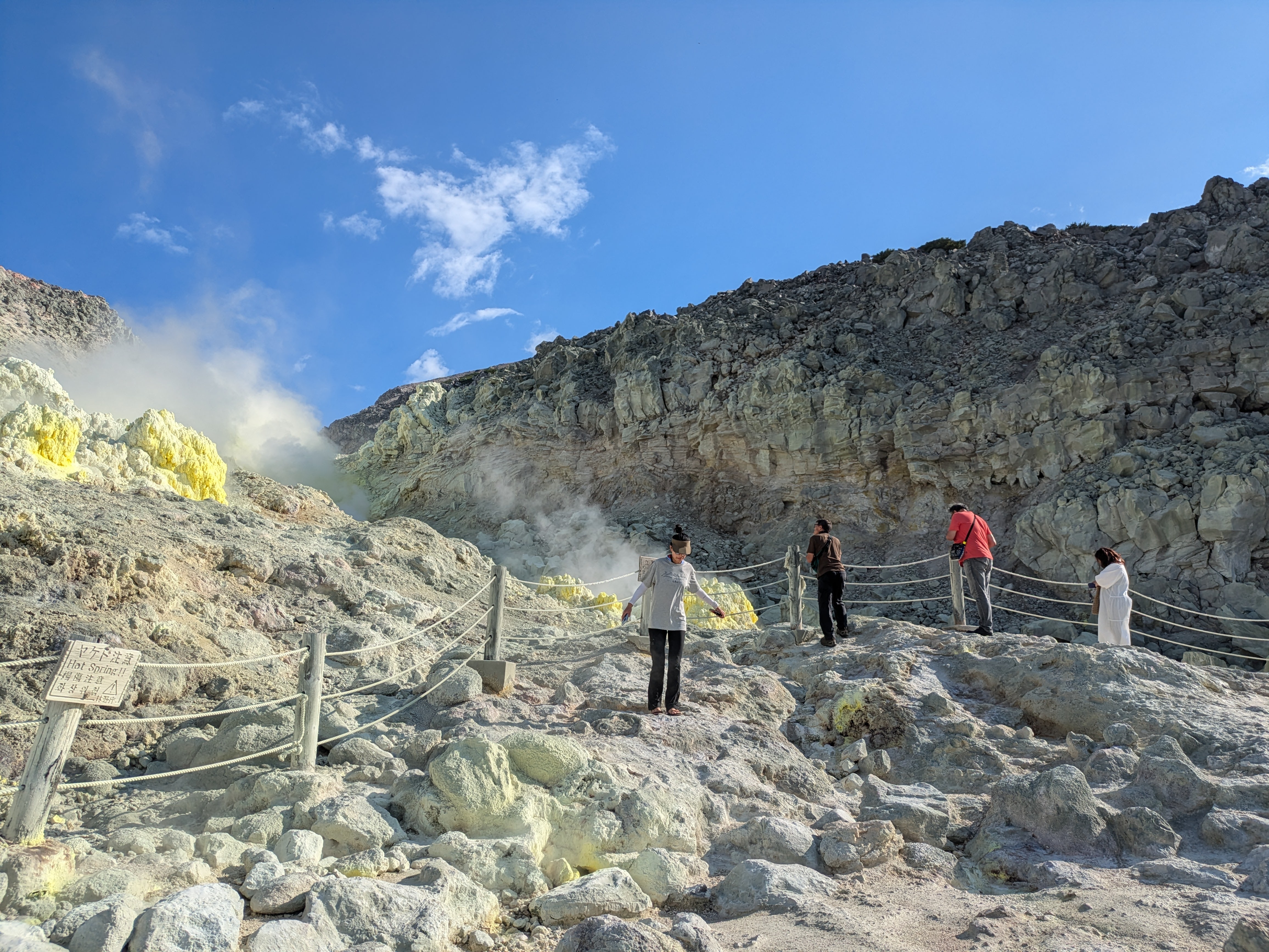 Four hikers look at the steaming vents of Mt. Io, Hokkaido, from behind a safety fence. The terrain is very rocky. Some of the rocks beyond the fence are a very bright yellow on account of the sulphur.