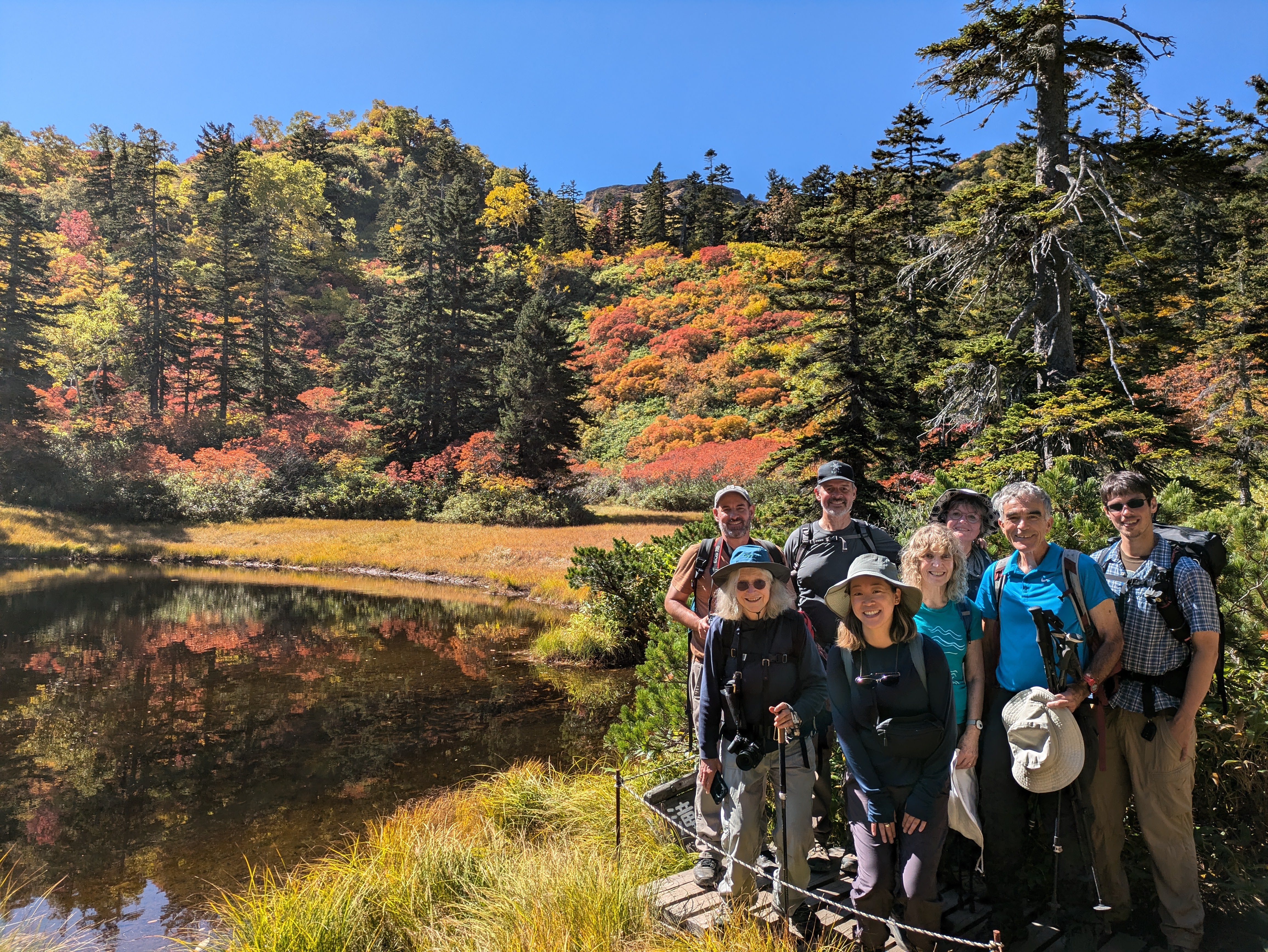 A group of hikers smile at the camera at Daisetsu Kogen, Hokkaido. Behind them is an alpine tarn (a pond) and plants and trees in autumn foliage on the other side. It is a beautifully sunny day.