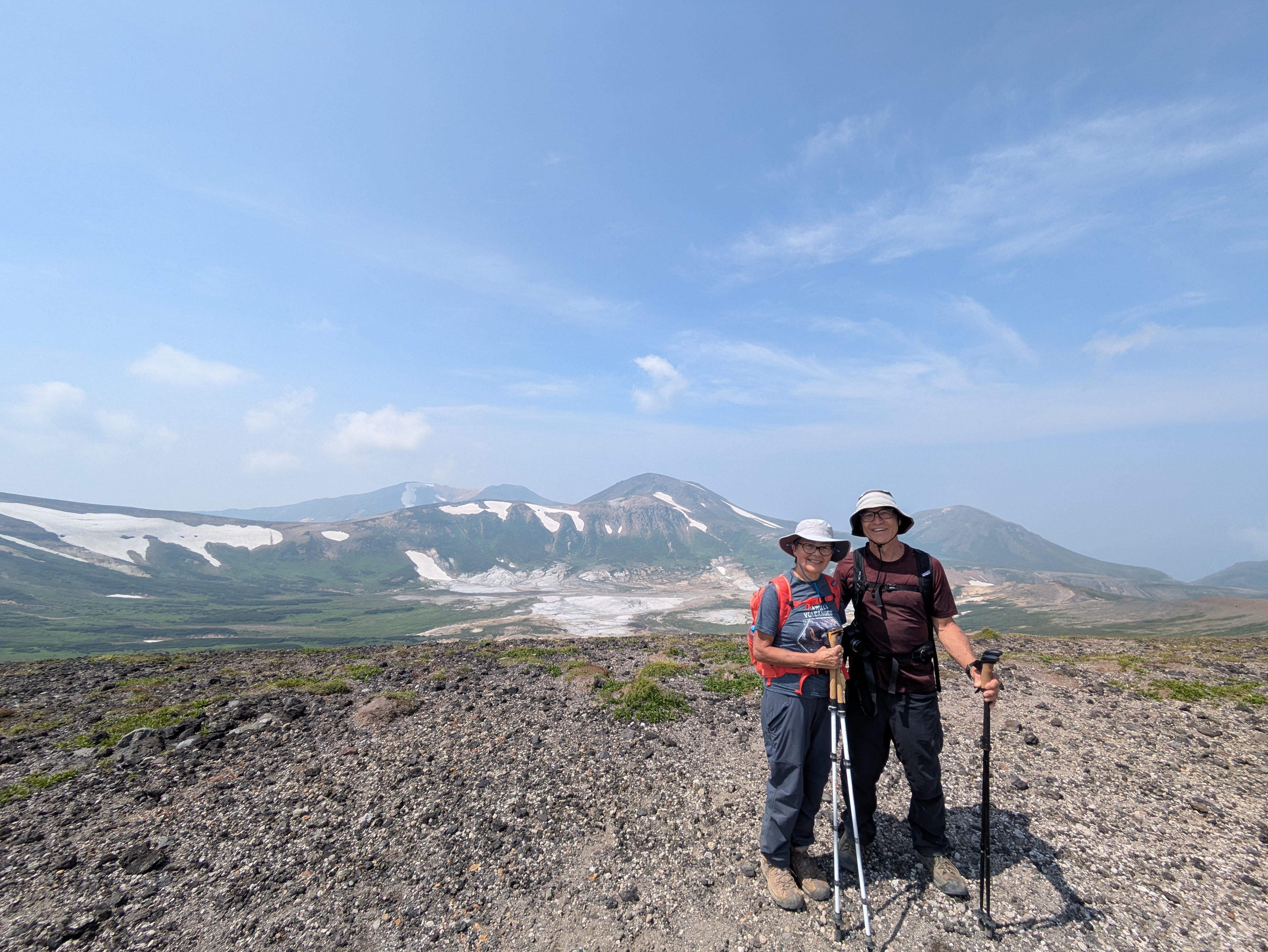 A couple put their arms around each other and smile as they stand on a plateau in the mountains. A large basin is visible behind them and mountain peaks in the distance. It is a very sunny day.