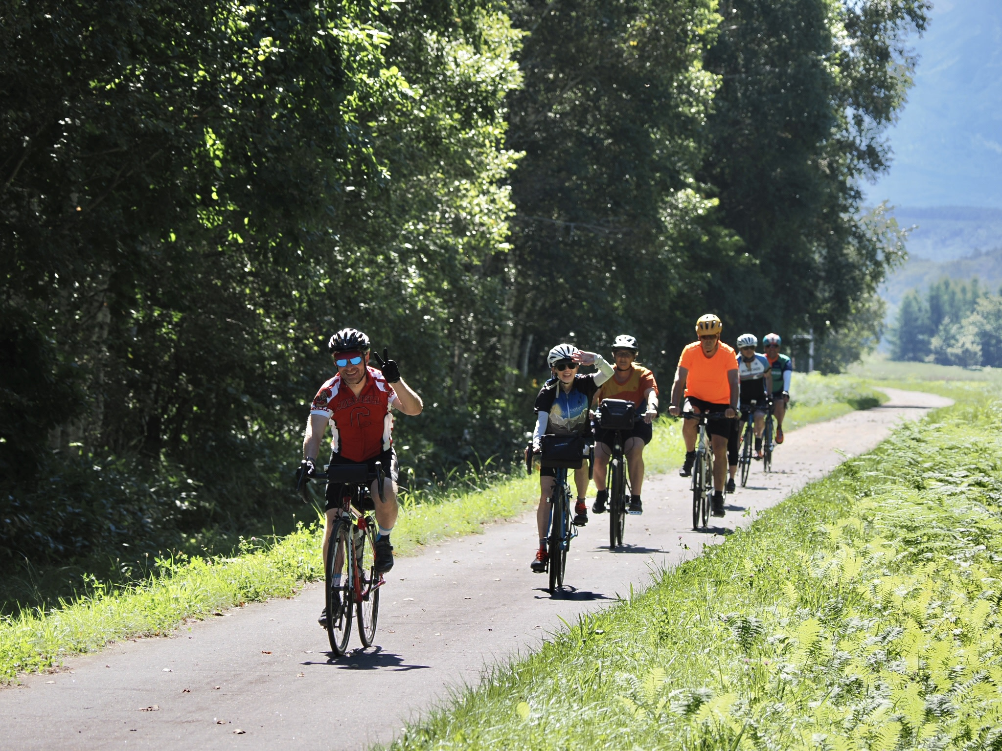 A group of cyclists ride along a cycling road towards the camera. The man at the front of the group is giving the camera a "peace" or "victory" sign (holding up the index and forefinger in a V-shape). The woman cycling behind him is giving the camera a salute. There is a forest to their right (our left).