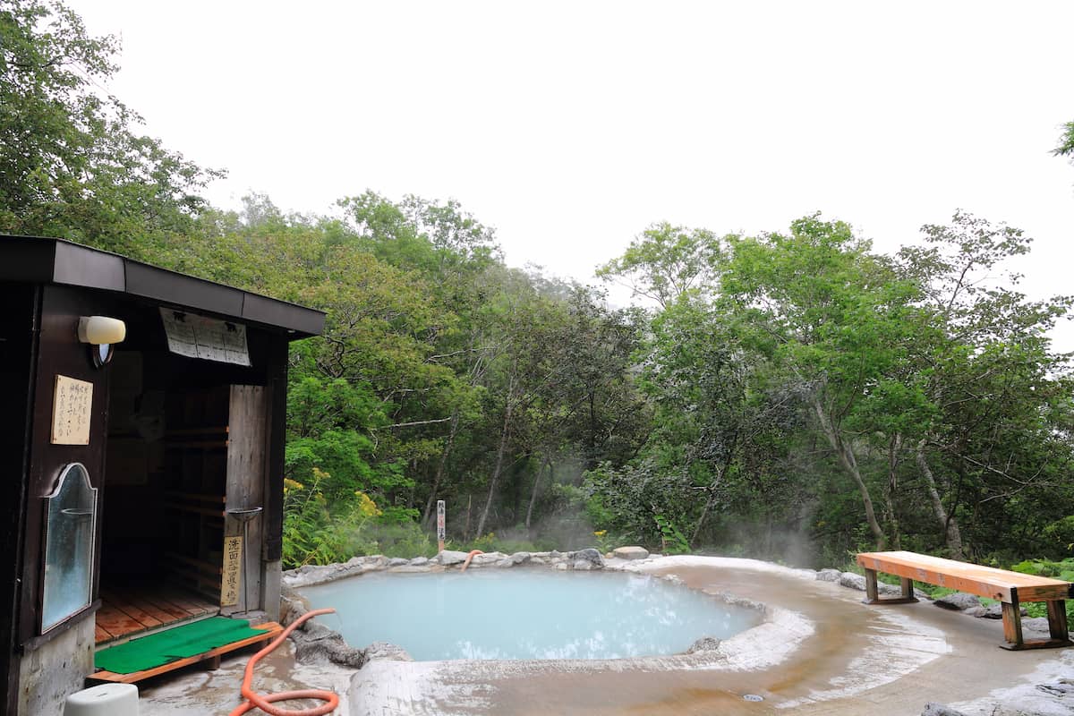 A milky white "rotenburo" hot-spring pool is surrounded by trees. A small changing room and bench sit either side of the onsen pool.
