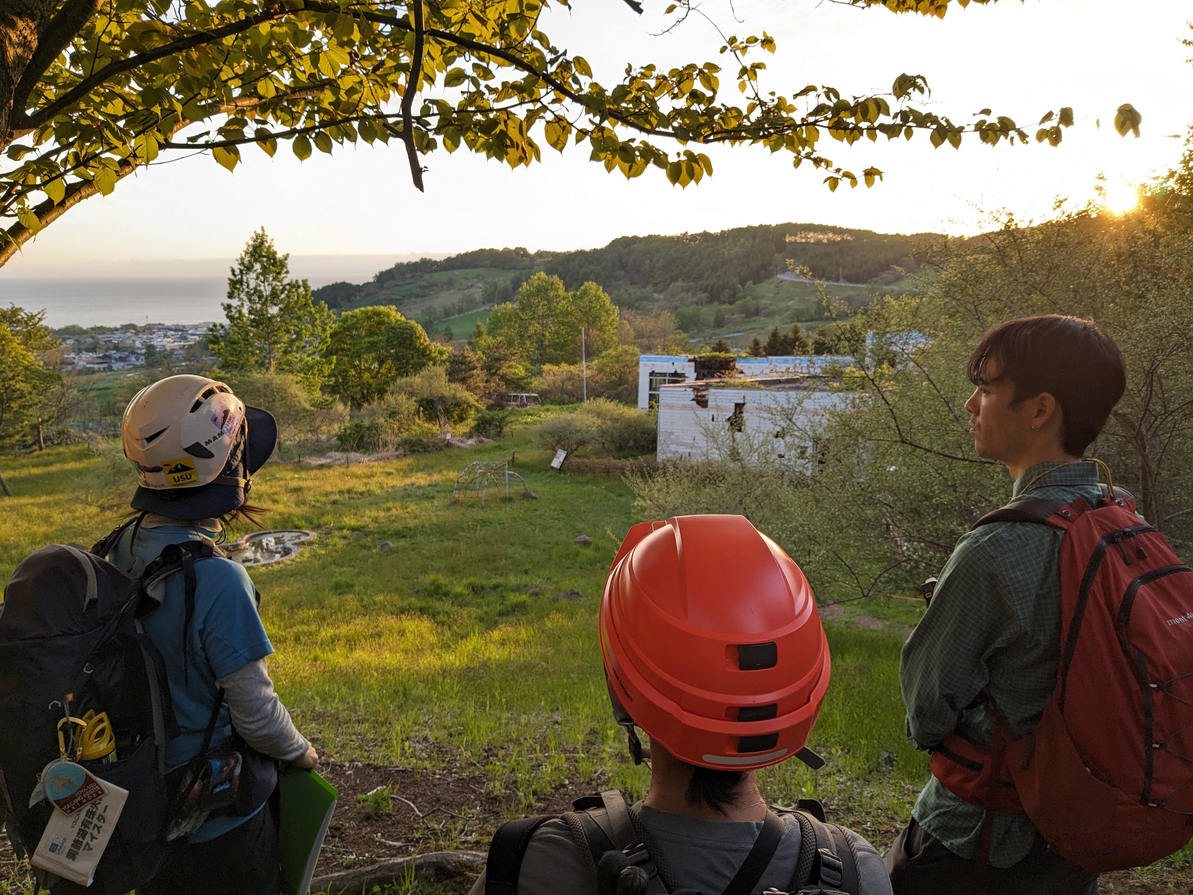 Three people look out over the ruins of a kindergarten destroyed by a 2000 volcanic eruption. Its walls are pockmarked with holes where it was struck by rocks. Volcanic rocks lie scattered about the grounds.