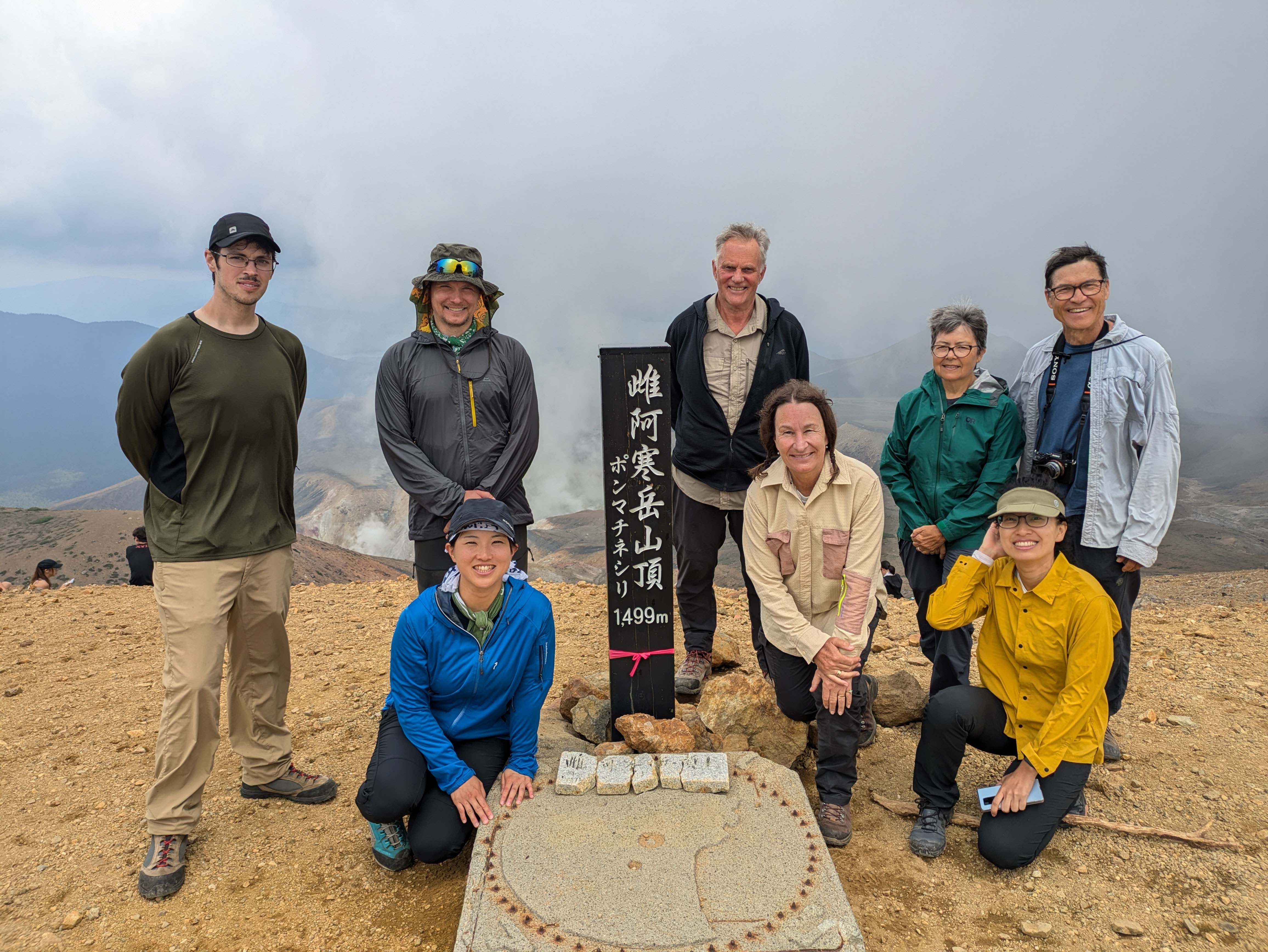 A group of hikers at the summit of a mountain. A pole in Japanese reads "Mt. Meakan Summit, 1499m". There are clouds and fog behind them.