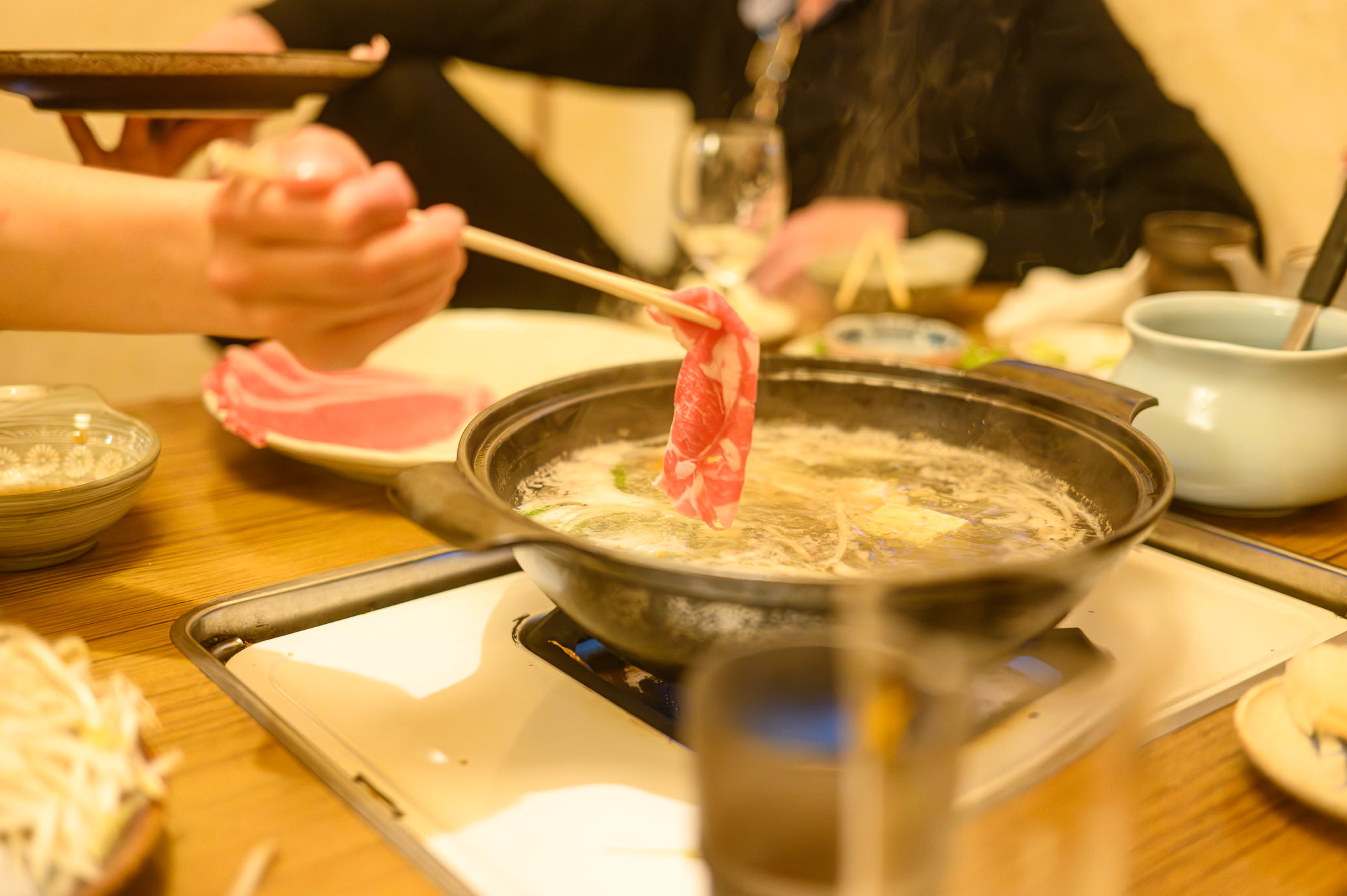 A hand holding a pair of chopsticks dips a thin slice of lamb into a pot of boiling broth.