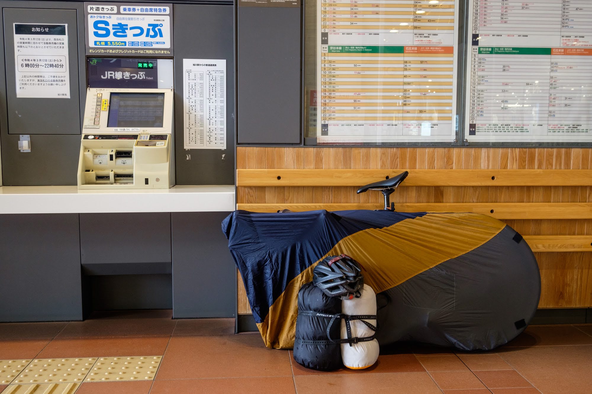 A bicycle is packed into a soft "rinko" bag for travel on the train. The saddle is poking out of the top of the bag and other luggage is stored in dry bags. The gear is leaning on a wall next to a ticket machine.