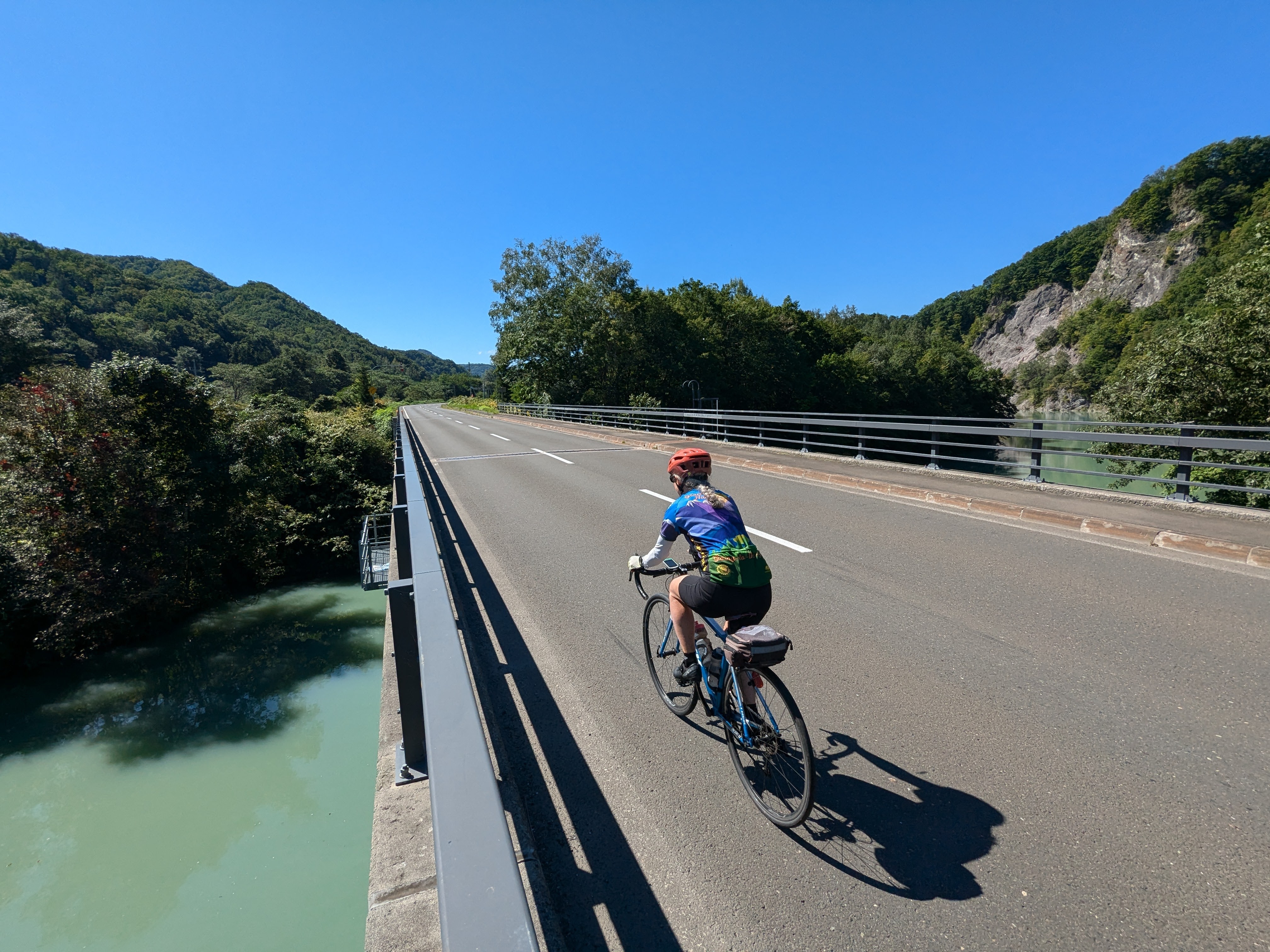 A cyclist crosses a wide river underneath a bridge. It's a wonderfully sunny day.