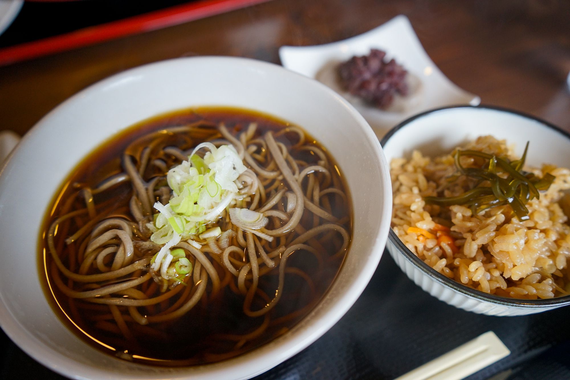 A close up of a bowl of soba noodles in broth, topped with green onions. There is a bowl of rice next to it.