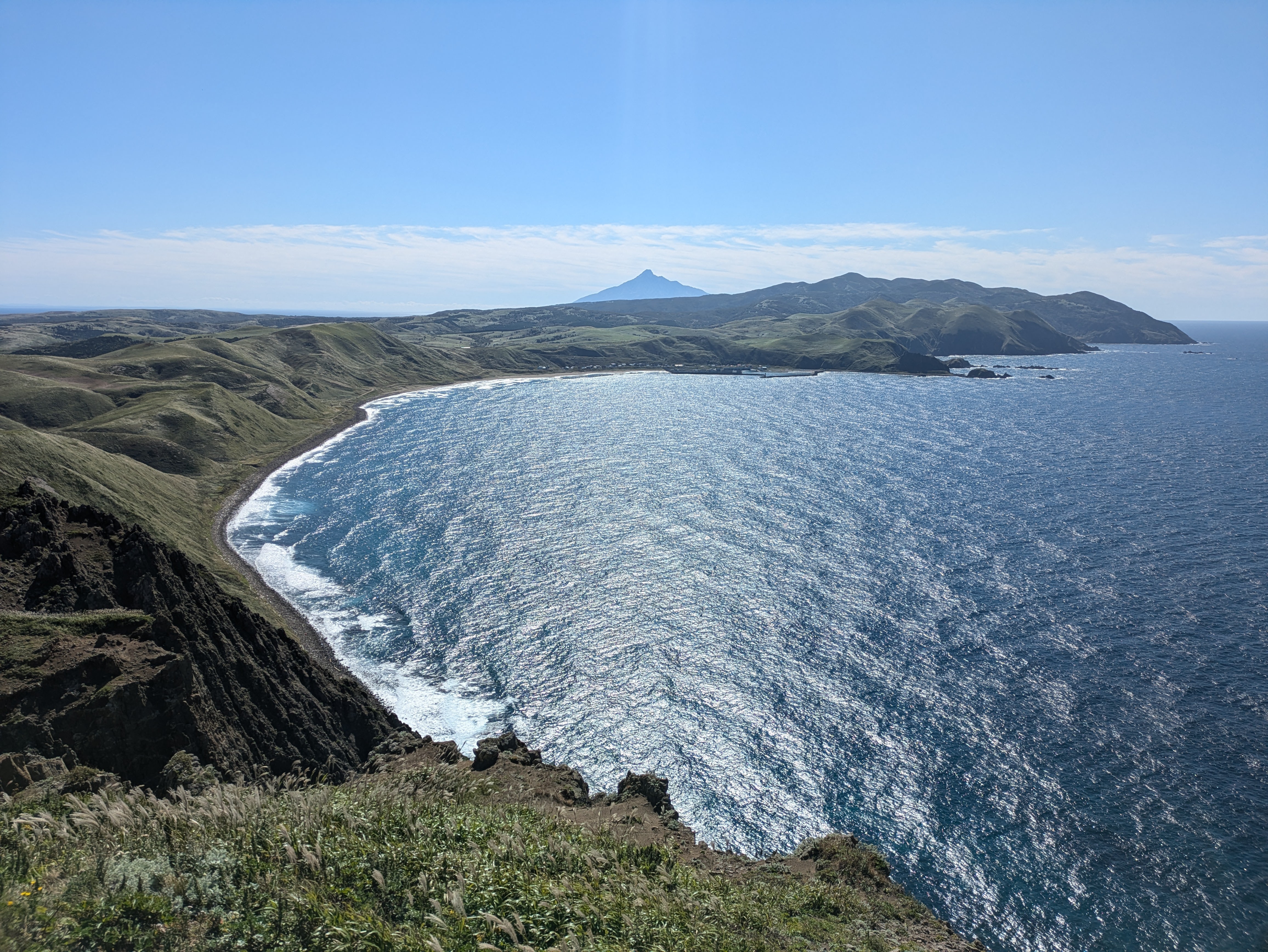 A photo of the view from the Misaki Meguri hiking course on Rebun Island. There is a beautiful view of an ocean bay far below and another cape in the distance. The summit of Mt. Rishiri is visible from behind the cape. It is a beautiful, sunny day.