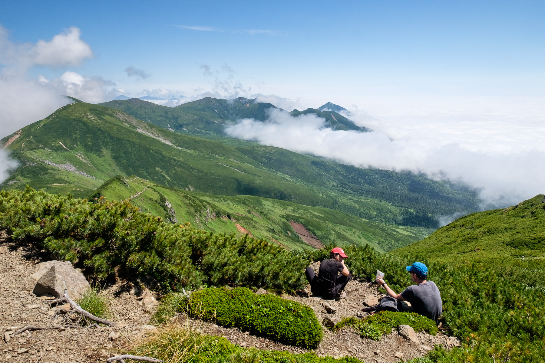 Two hikers share lunch at the summit of Mt. Furano. The mountain ridge stretches into the distance, surrounded by a sea of cloud