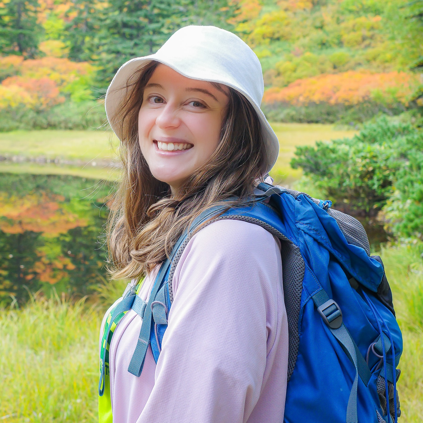 A female hiker wearing a cute white hat beams into the camera. In the background the first autumn colours in the Daisetsuzan are reflected in a still pond at Kogen Onsen.