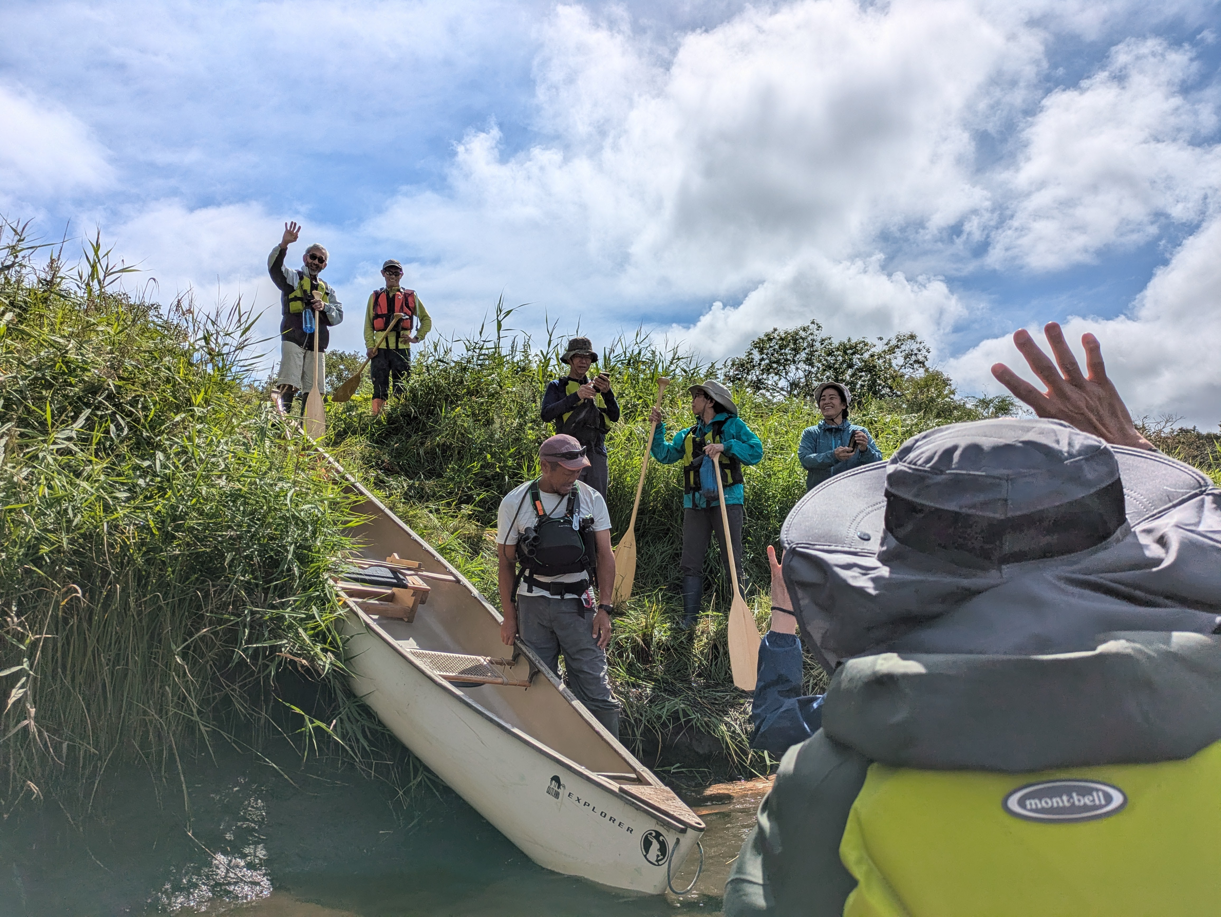 A group of people stand on the grassy shoreline of a river, holding oars. In the foreground is someone in a canoe, waving to the others on the shore. A man is preparing a canoe to go into the water in the midground.