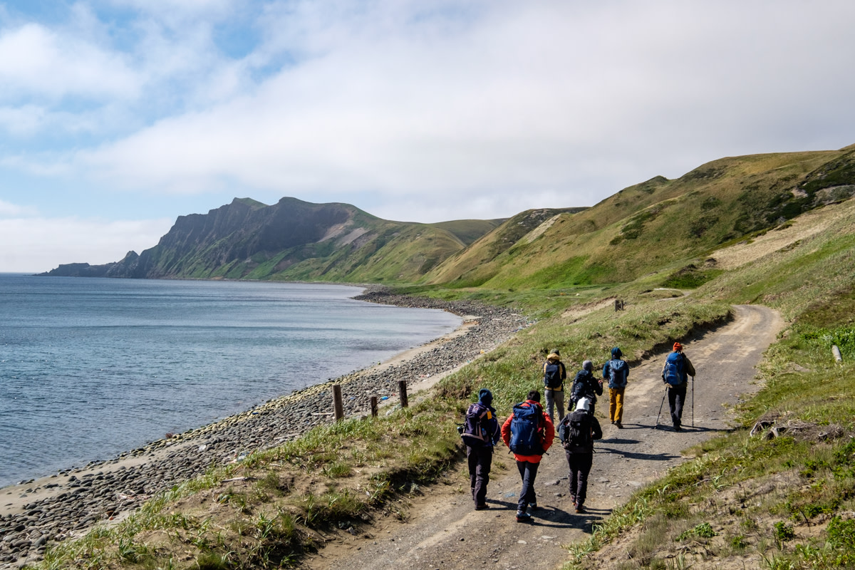 A group of hikers walks along the shoreline of Rebun island, with the dramatic cliffs of Cape Gorota in the background