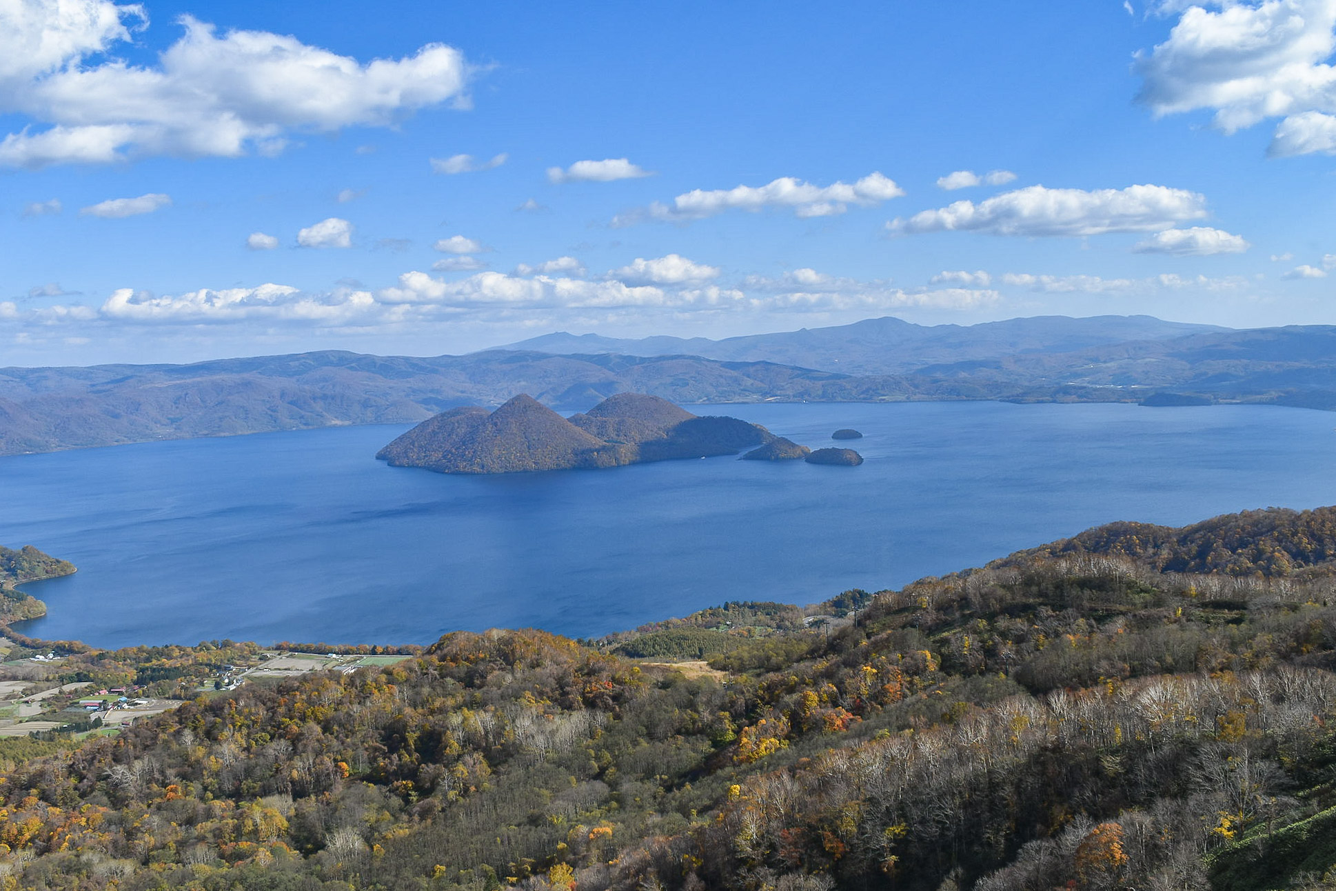 A view of Lake Toya from up high. Most of the lake is visible. Nakajima Island is visible in the middle of the lake. It is a bright, sunny day.