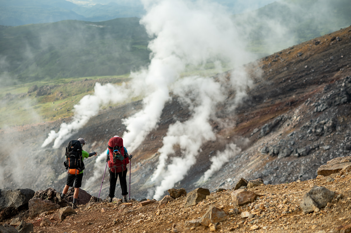 Two hikers stand on a rocky ridge and look out over white plumes of steam rising from volcanic vents on Mt Asahidake