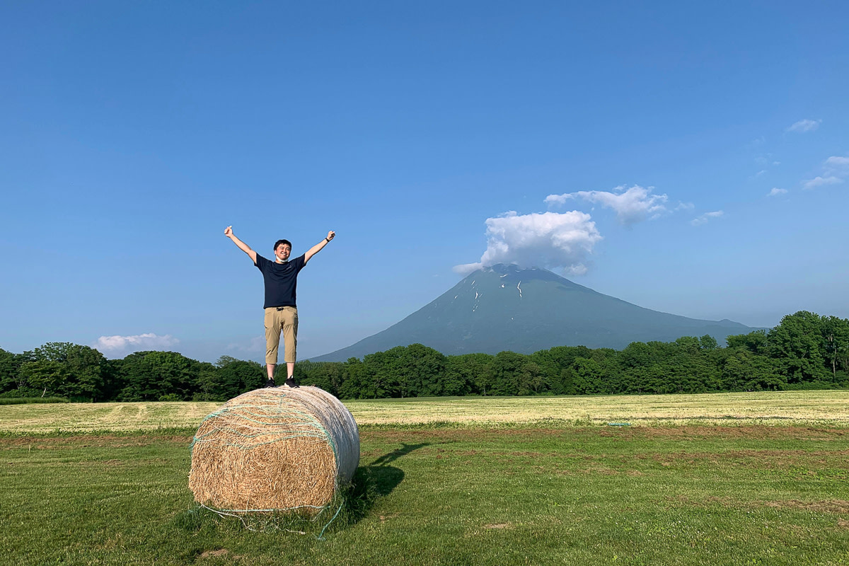A traveler poses on top of a hay bale with Mount Yotei in the background