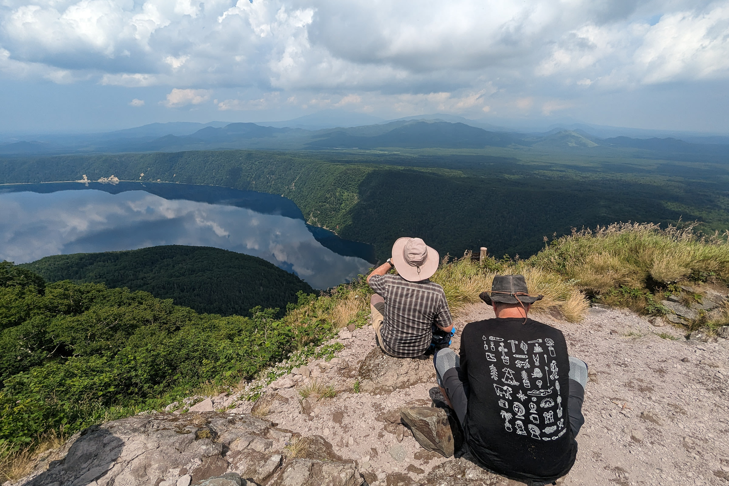 A group of hikers take a break on a mountain, looking out over a partial view of Lake Mashu. It is a still day and the water's surface is very reflective.