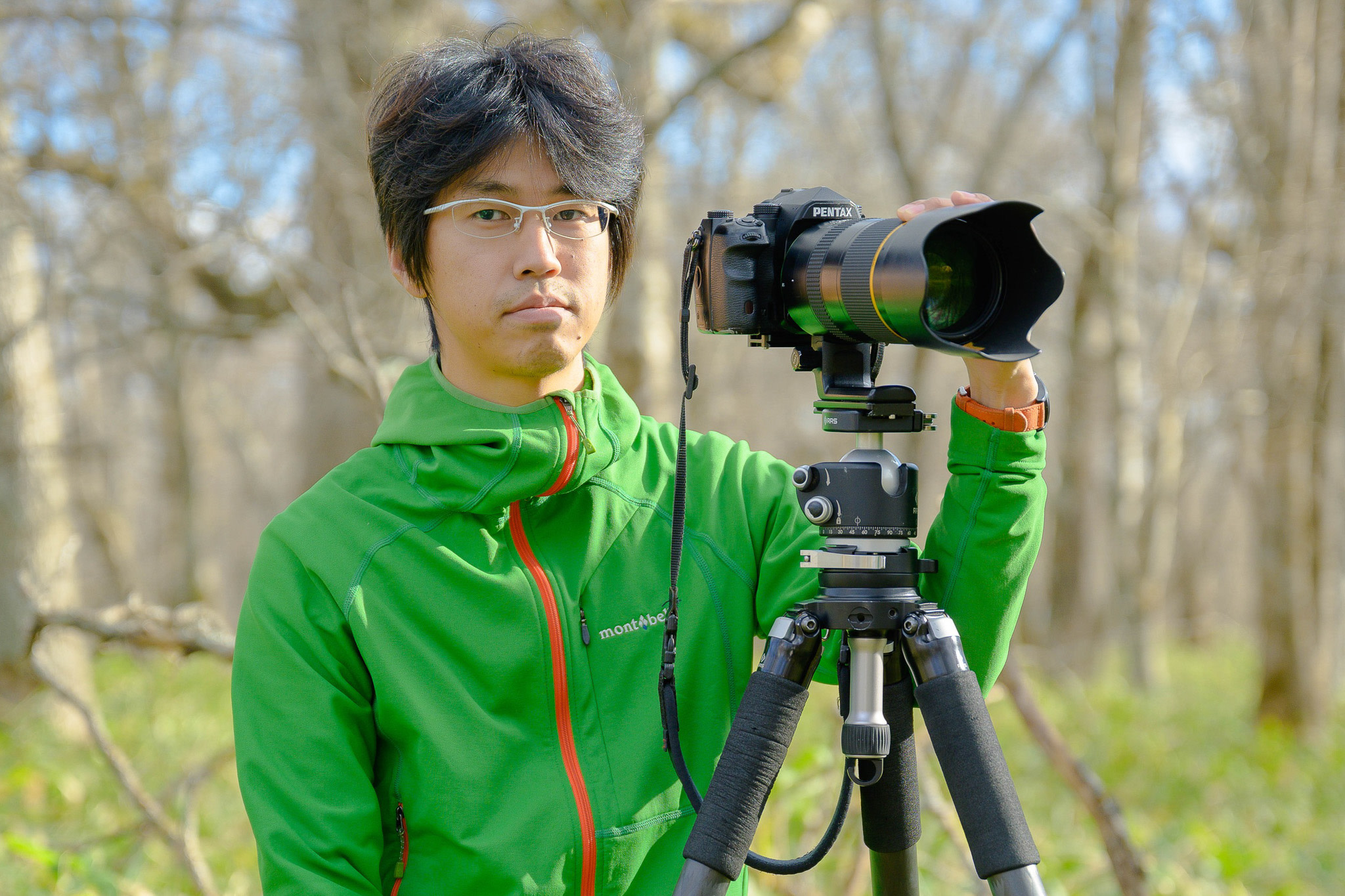Yuto Hirasawa, an adventure Hokkaido guide, smiles at the camera. He is stood behind a camera mounted on a tripod. His hand rests on a large zoom lens.