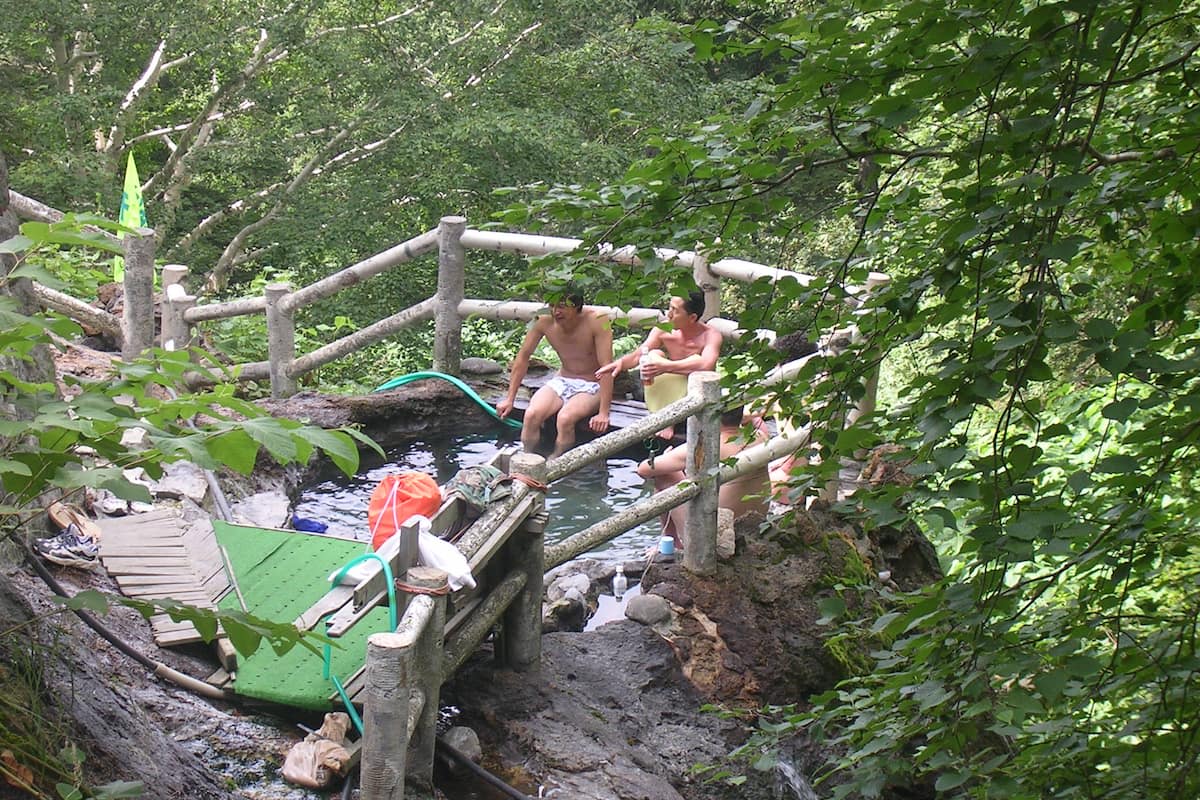 4 men sit around a wild onsen hot spring pool. The pool is surrounded by green leaves and birch trees.