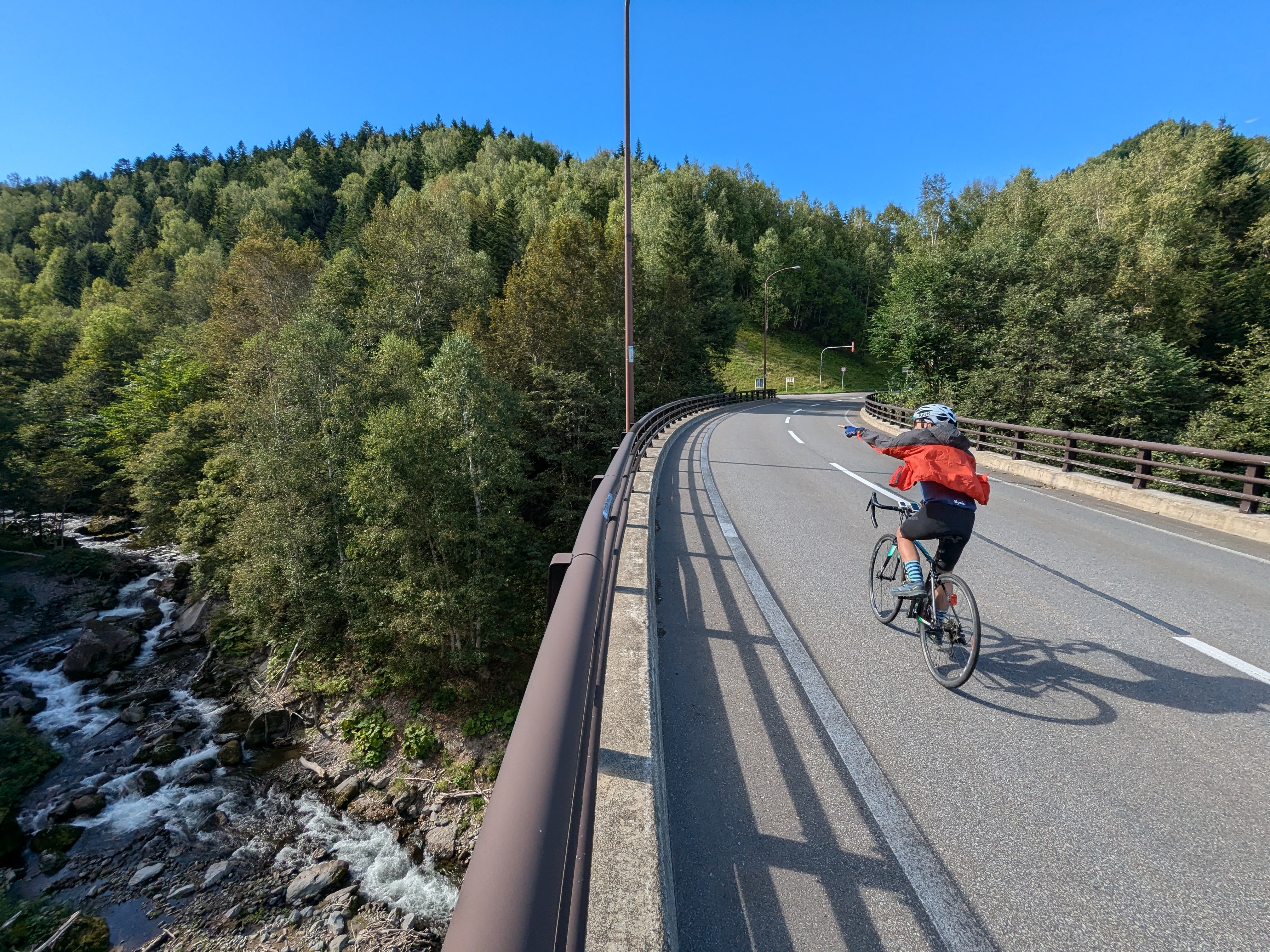 A cyclist crosses a bridge over a river. They are pointing out towards the river. It is a beautiful sunny day.