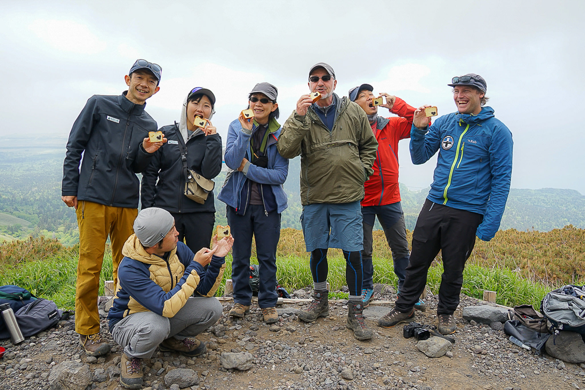 A group of hikers at the summit of a mountain all hold up a piece of cake to show the camera. The cake has a heart shape running through it.