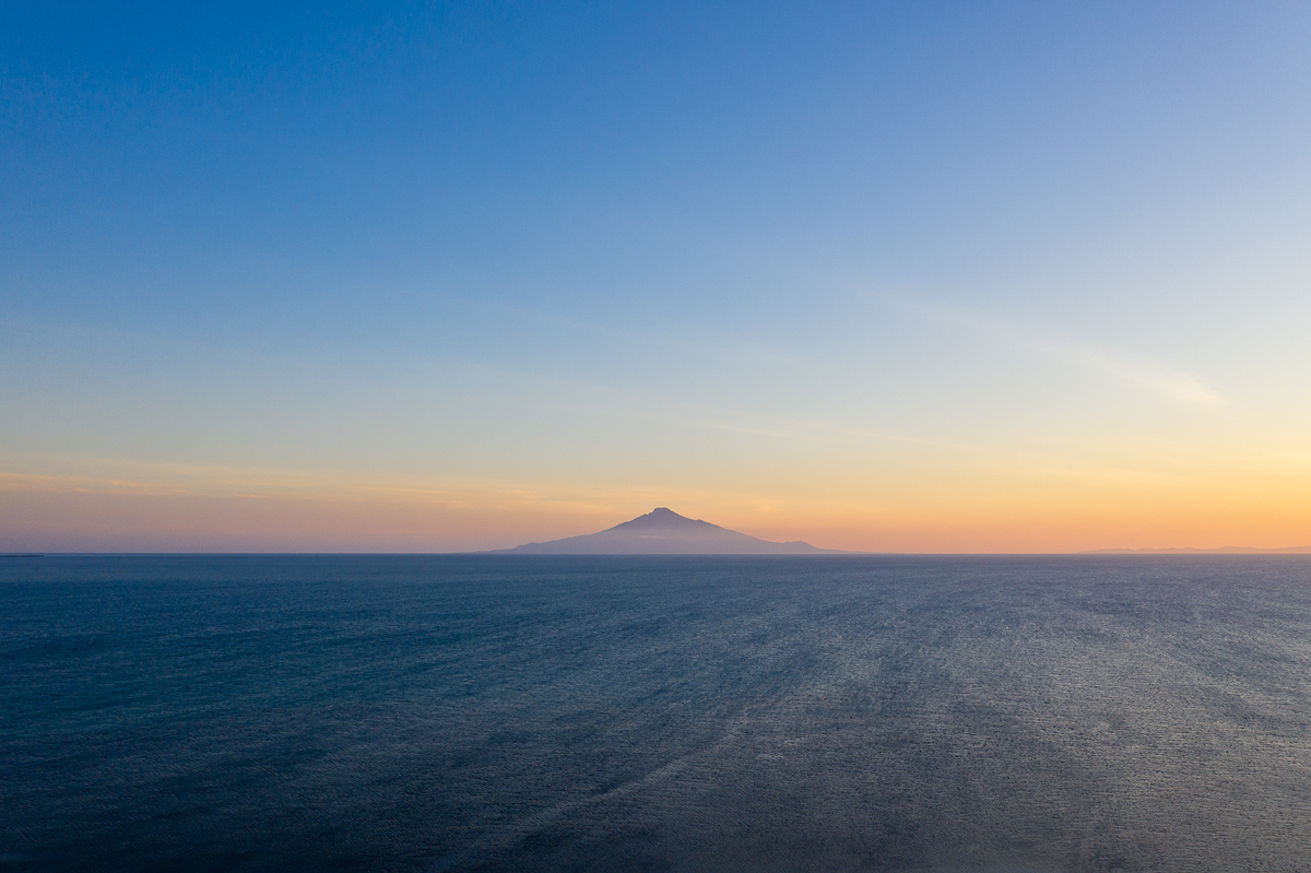 Mt Rishiri is seen floating on the horizon at sunsen. The sky has an orange strip along the horizon, graduating to deep blue.