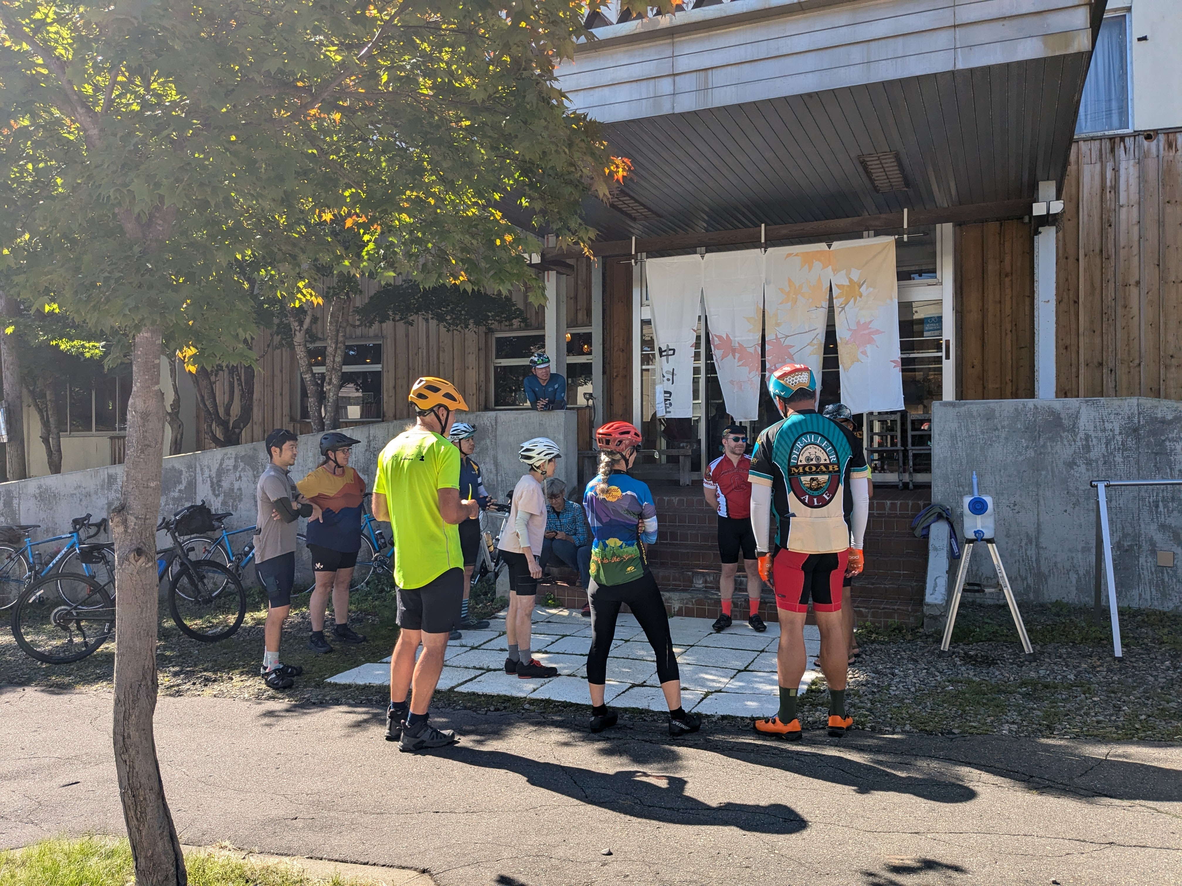 A group of cyclists stand at the entrance to a Japanese inn. The inn has cloth curtains hung in front of the door with the image of autumn leaves to celebrate the autumn season.