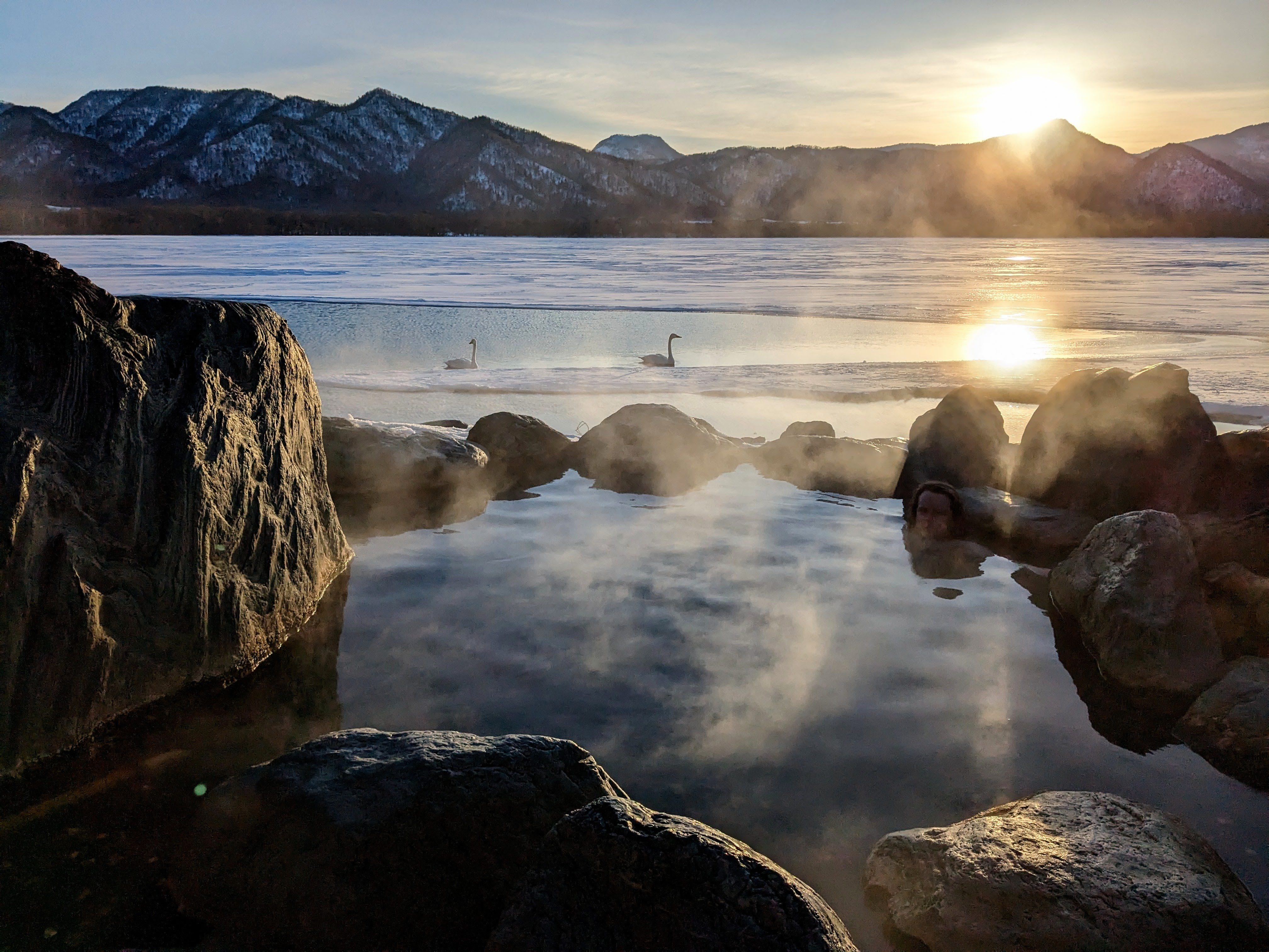 A bather soaks in a wild onsen on the shore of Lake Kussharo. There is ice on the surface of the lake and a pair of Whooper Swans float in an open patch of water. It is sunset and the suns rays are backlighting the steam rising off the hot spring pool.