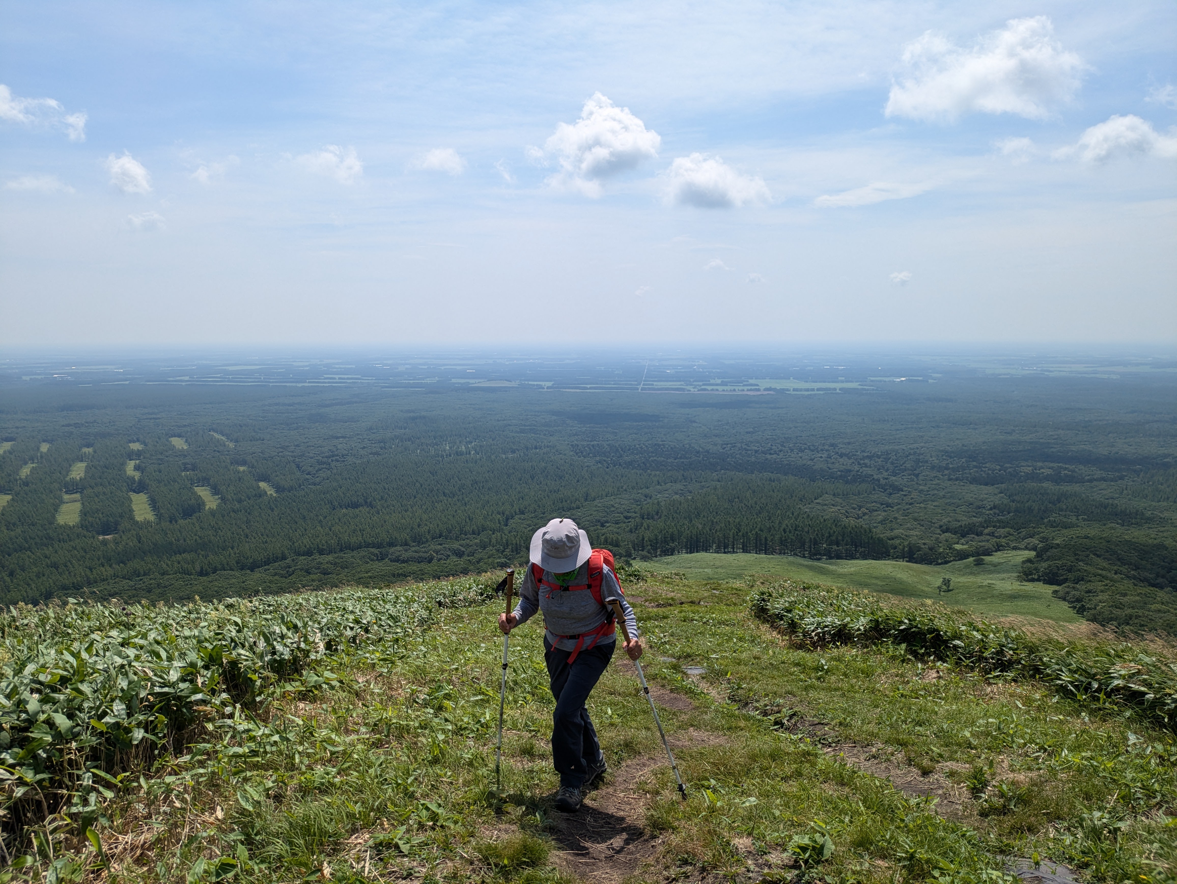 A woman hikes up a very steep, grassy hill. Behind her, there is a vast, flat plain in the distance.