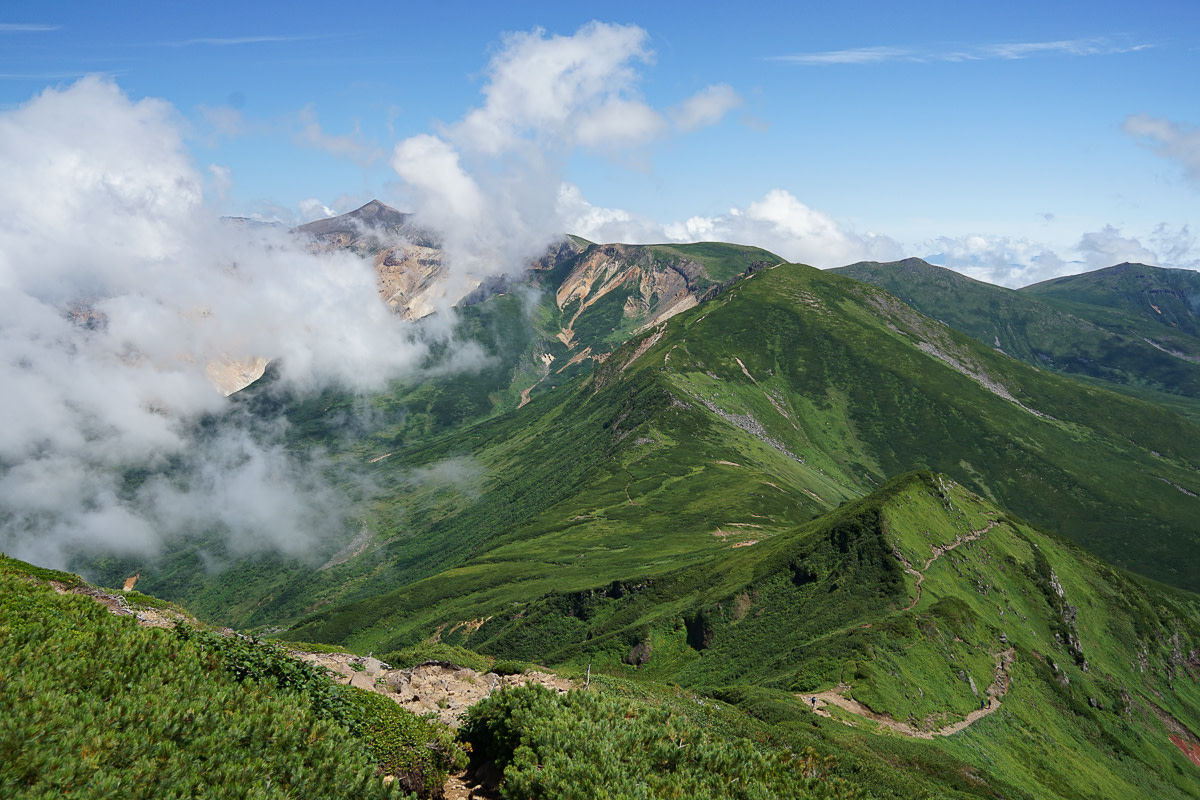 The green ridgeline of the Daisetsuzan mountains stretches away in the distance. The summit of Mt Tokachi is framed by clouds and the rest of the sky is blue.