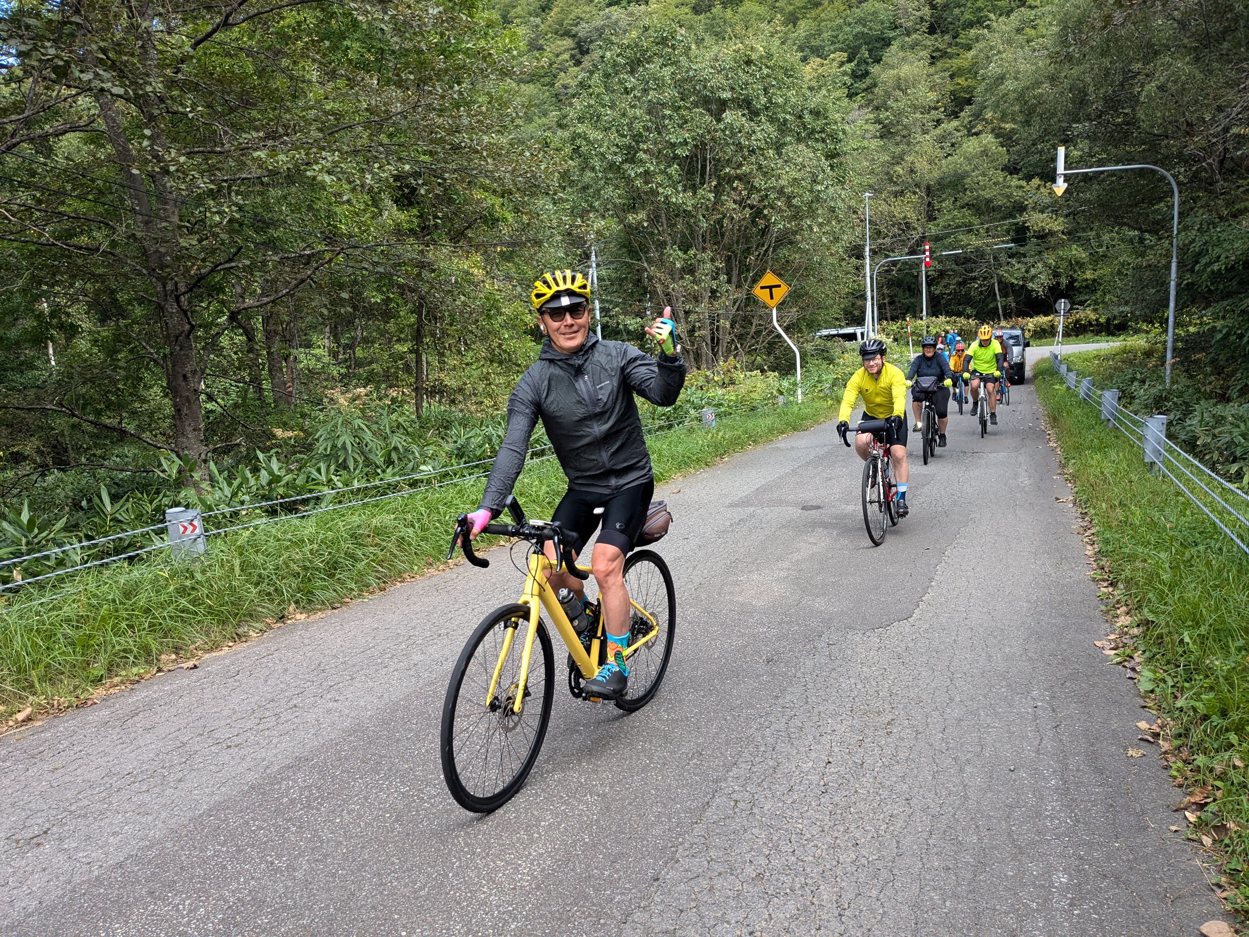 A group of four cyclists ride in single file on a narrow road. The cyclist at the front is giving the camera a "hang loose" gesture.