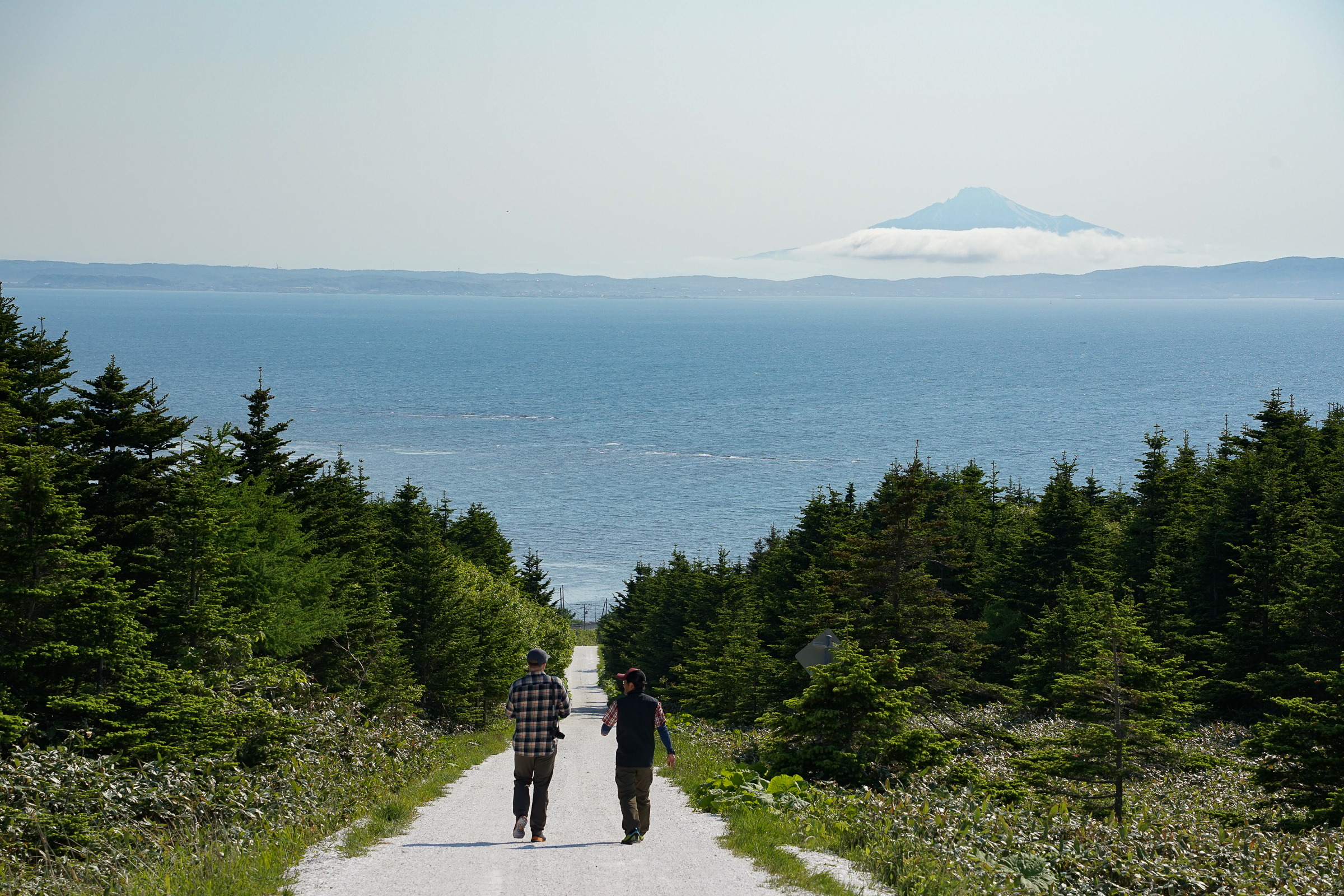 Two men walk down a white path leading to the sea, with Mt. Rishiri wreathed in cloud in the distance.