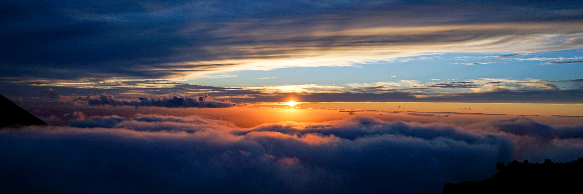 A spectacular sunset seen from high up in the Daisetsuzan mountains. The sun is just about to drop below the horizon, casting an orange light over clouds below the photographer. Dark clouds higher in the sky frame the sun.