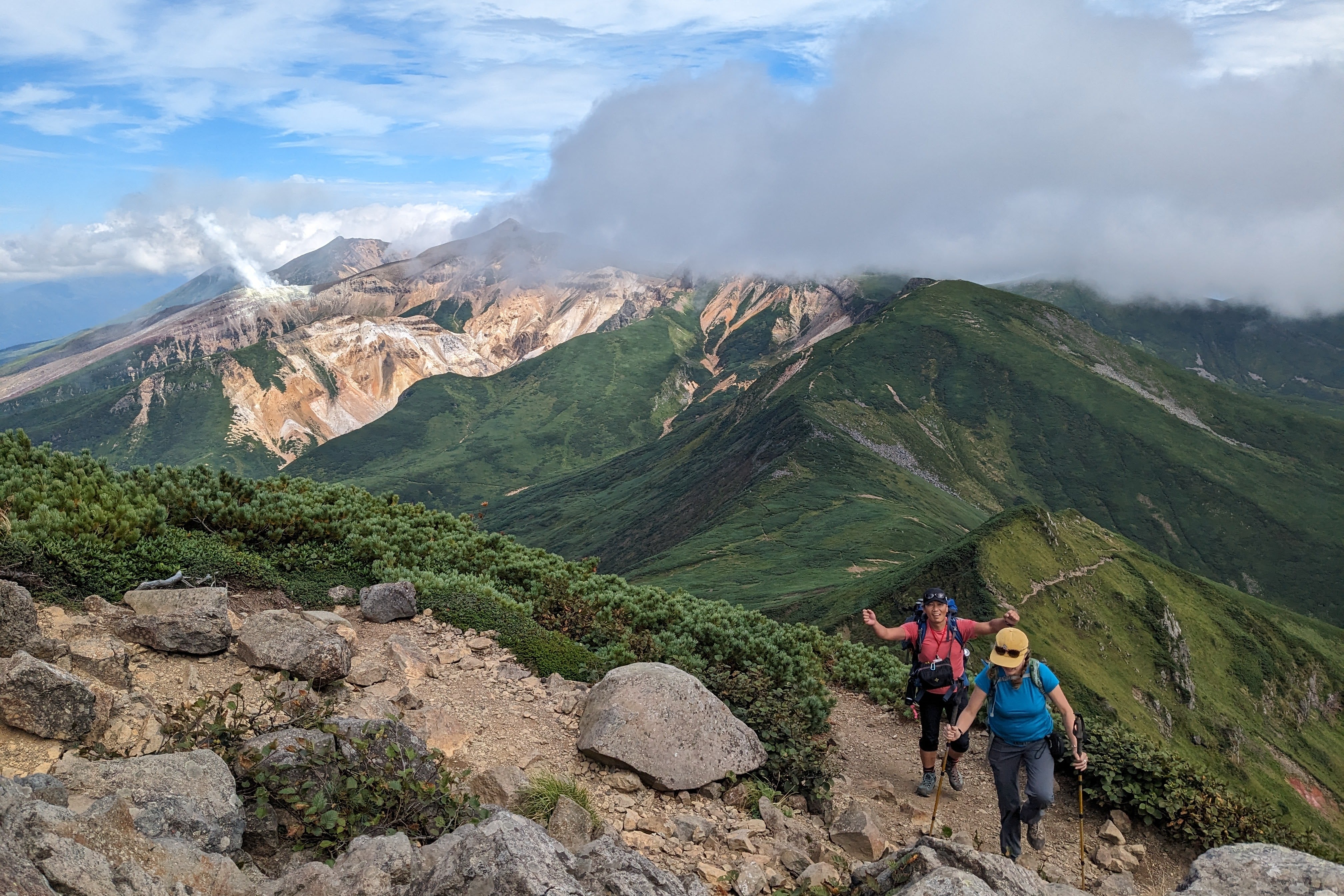 Two hikers make their way up a rocky hiking route on Mt. Furano, with green mountains in the background. One of them gives the camera a thumbs-up.