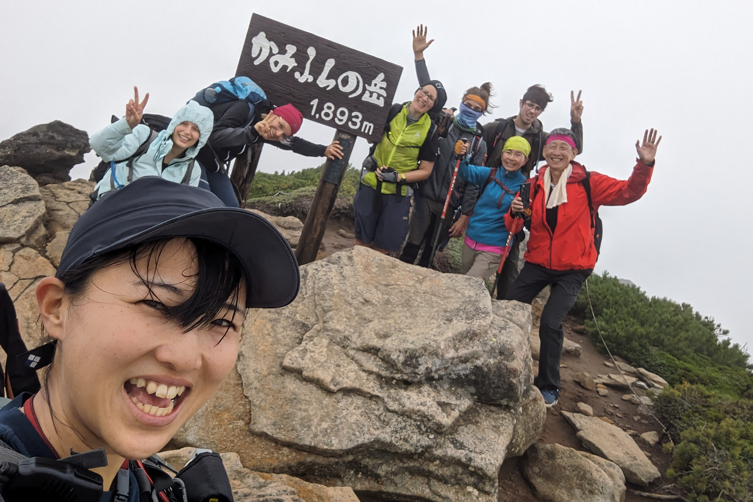 Adventure Hokkaido guide Yuka poses for a selfie with hikers atop a mountain. A sign in Japanese reads "Mt. Kamifurano - 1893m").