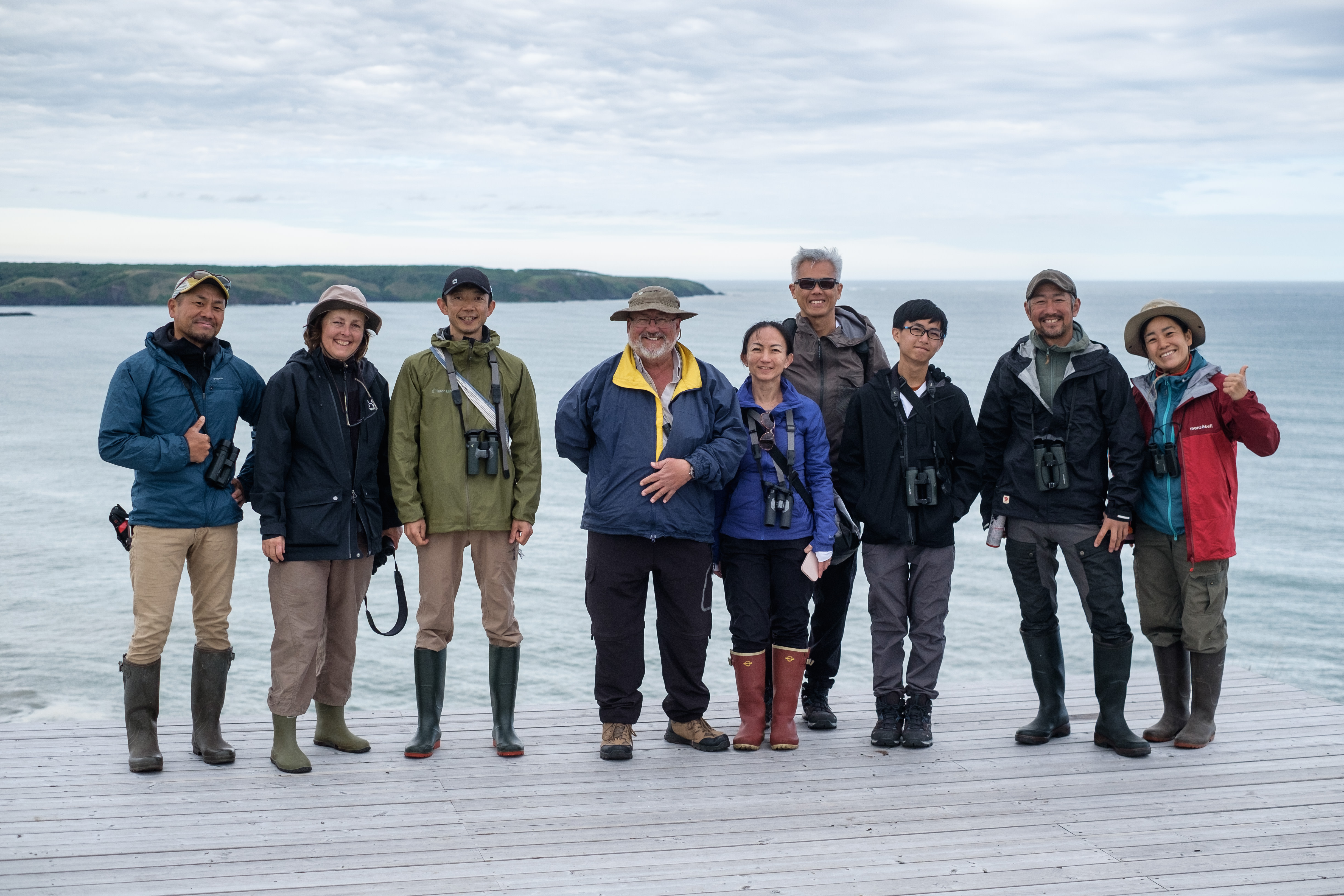 A group of tourists pose for a photo with local guides. The group stands on a wooden deck overlooking a grey sea with a windswept headland in the distance.