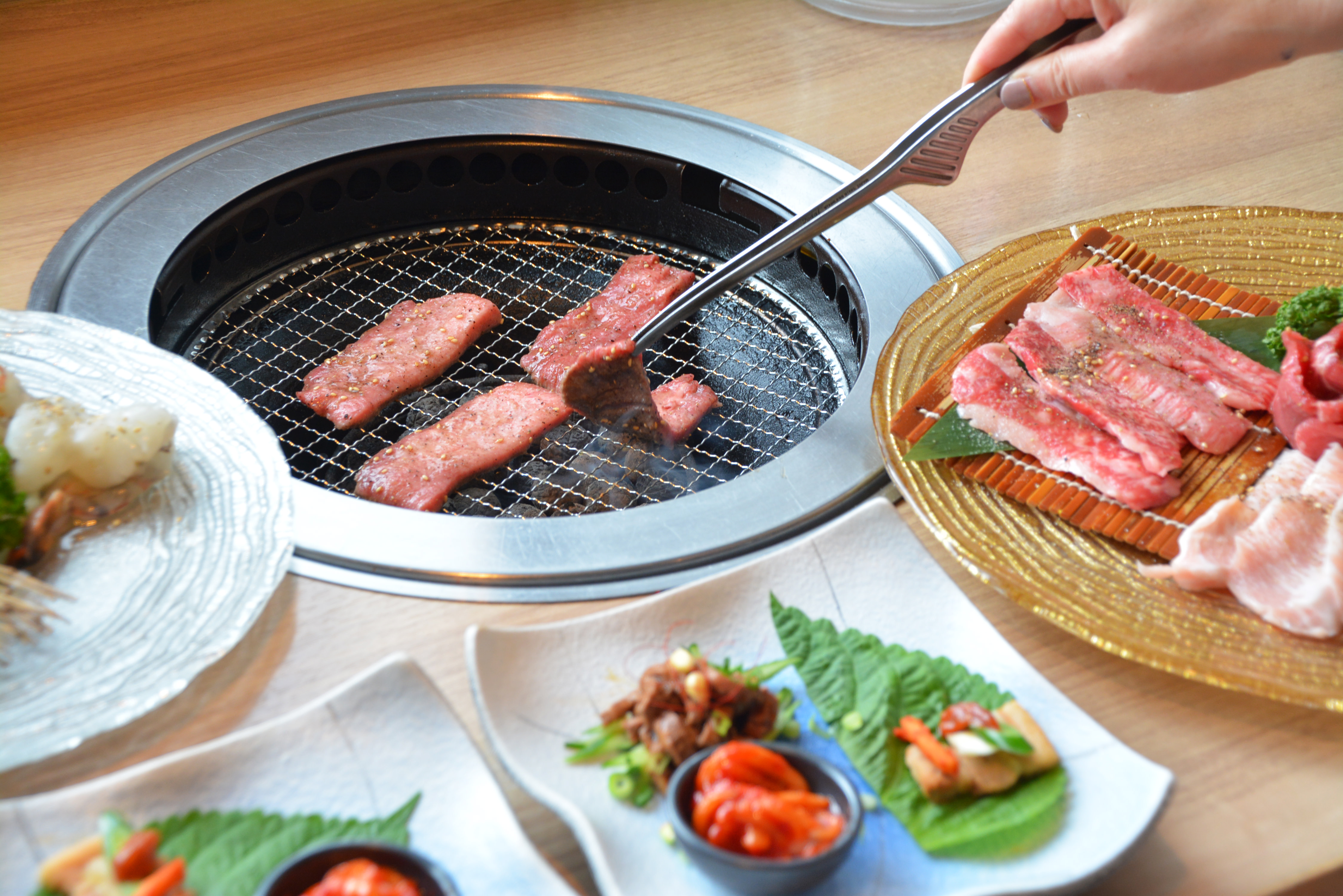 A hand uses metal tongs to grill meat on a meat grill. There are plates of meat and side dishes around the grill.