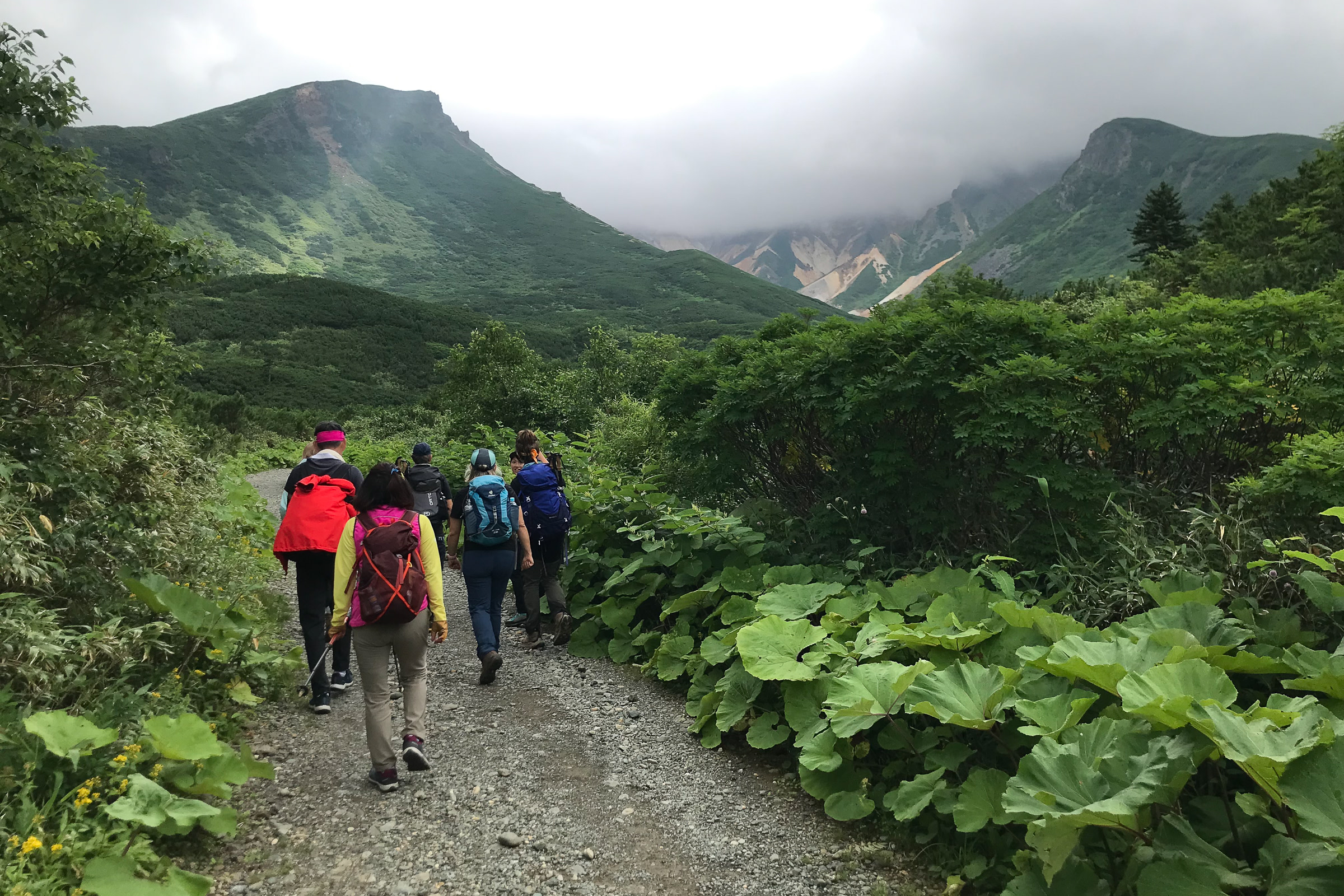 A group of hikers on a gravel path head into the mountains. A large volcanic crater is in the background. It is a cloudy day.