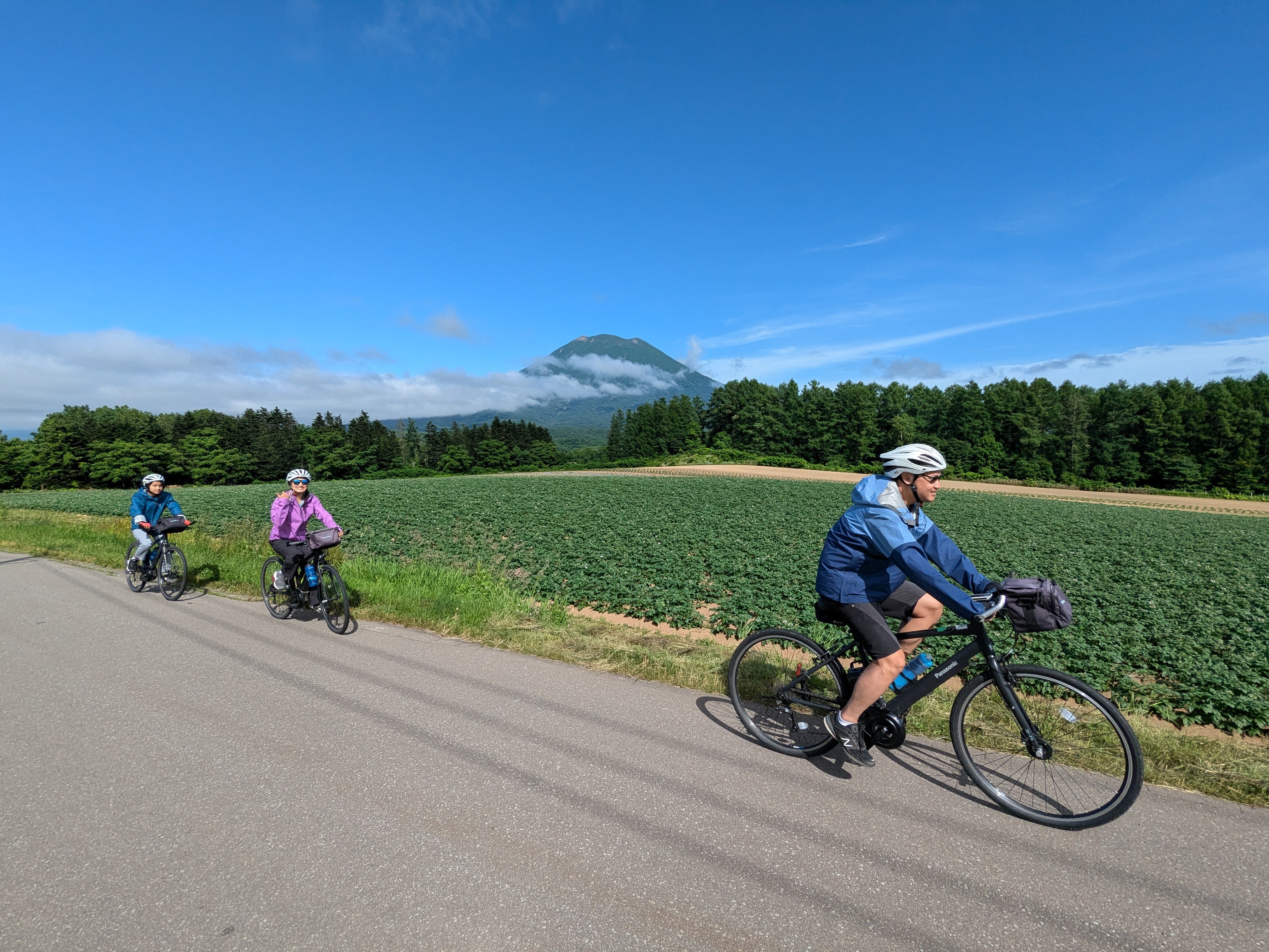 Three cyclists ride in a line past Mt. Yotei in Hokkaido. It is a beautifully sunny day. Mt. Yotei is partially obscured by clouds, giving it a mysterious look.