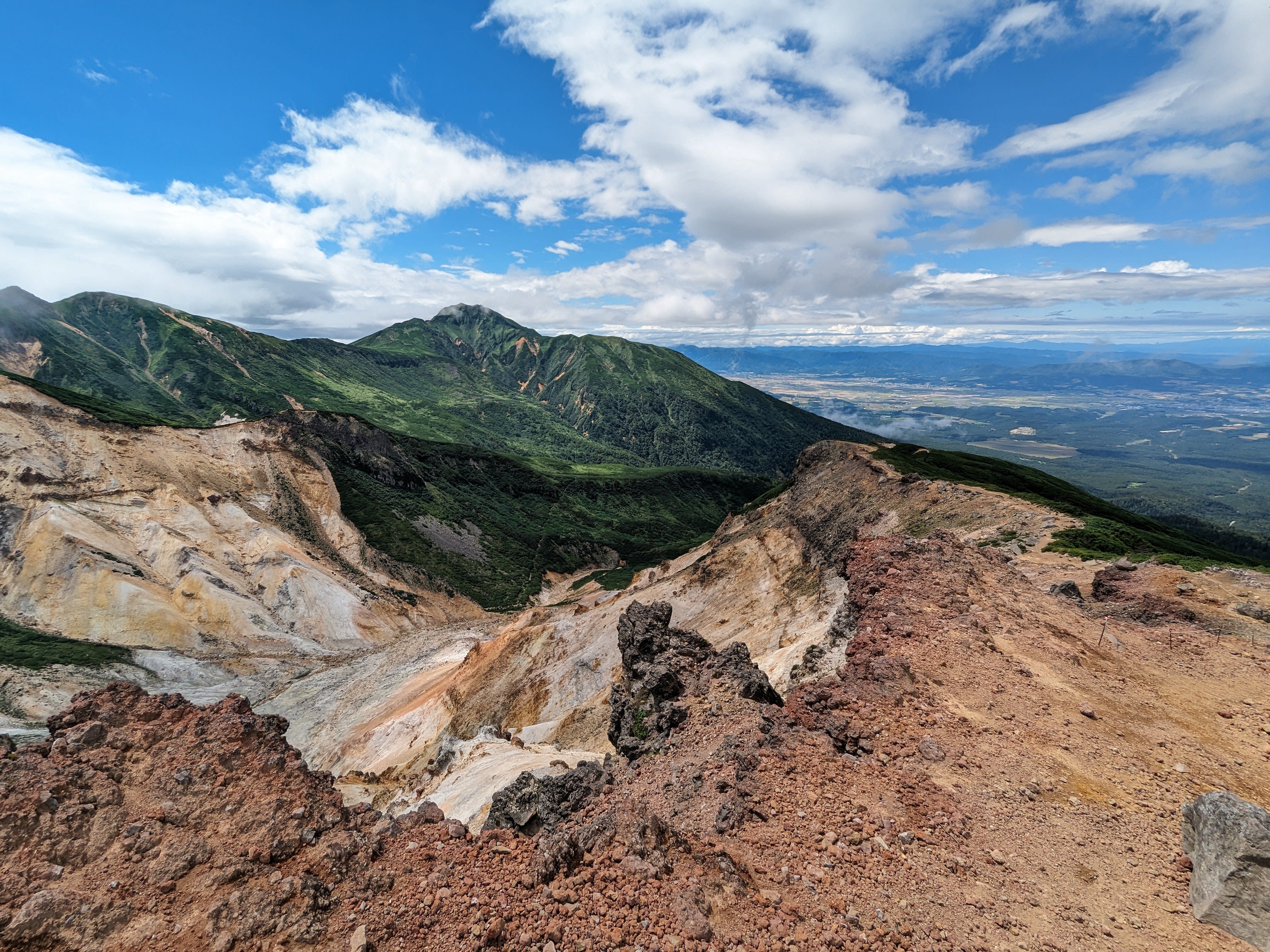 A green Mt. Furano is seen standing over the Furano plains. The photo is taken from Mt Sandan and looks out over the rocky landscape of Ansei Crater. The foreground ridge is also rocky, showing the volcanic nature of Daisetsuzan National Park.