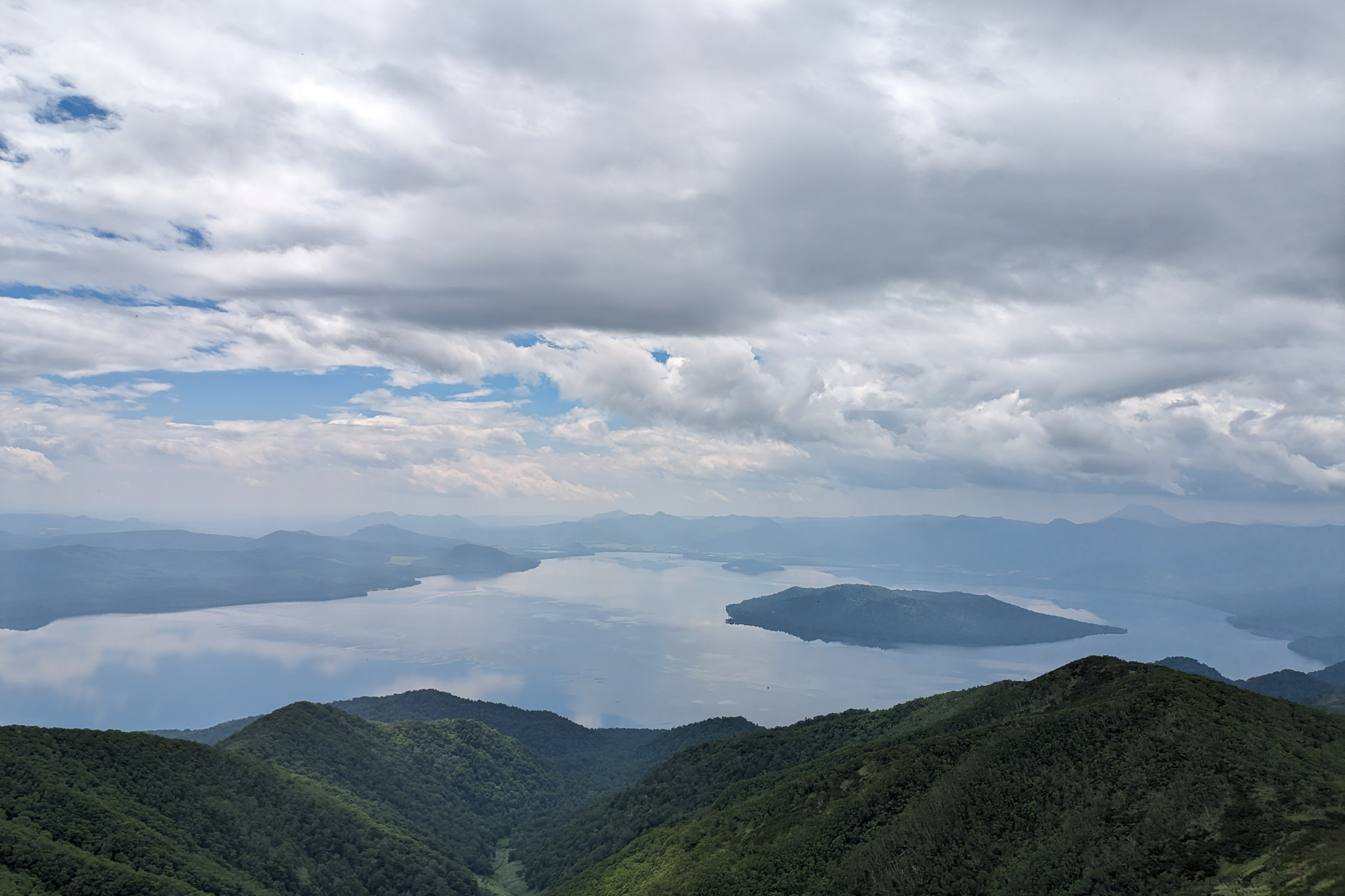 The view of Lake Kussharo from Mt. Mokoto. It is a partially cloudy day and an island is visible in the middle of the lake. 