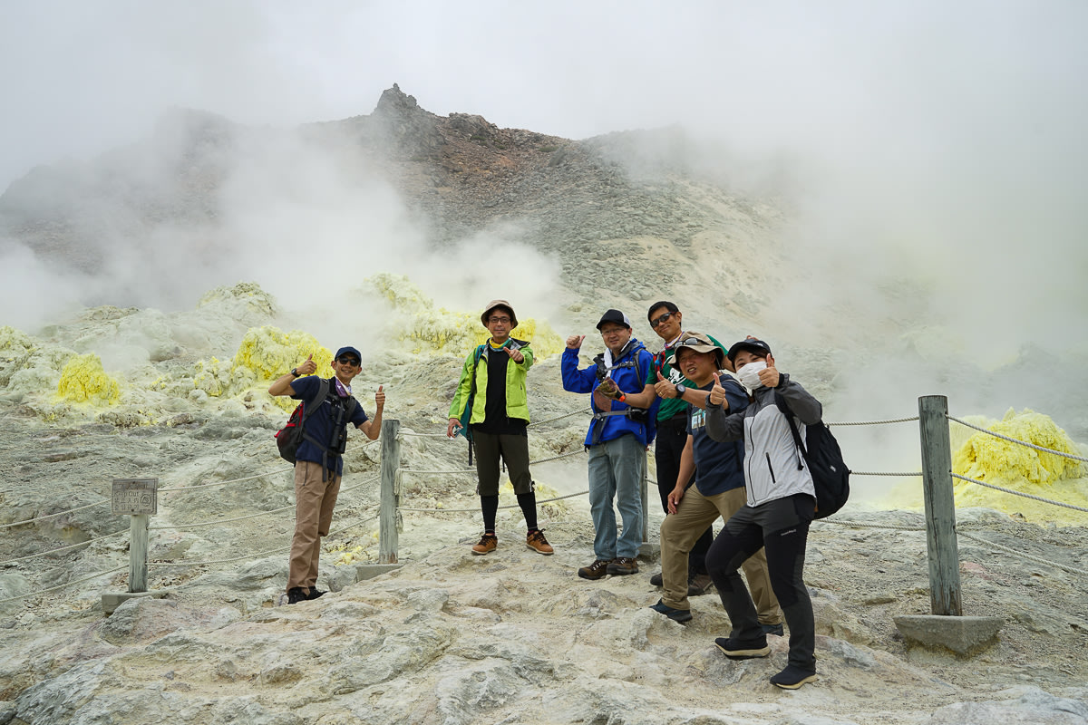 A group of guests on a guided adventure tour pose for a photo in front of steaming volcanic vents. The vents a ringed in yellow sulphur.