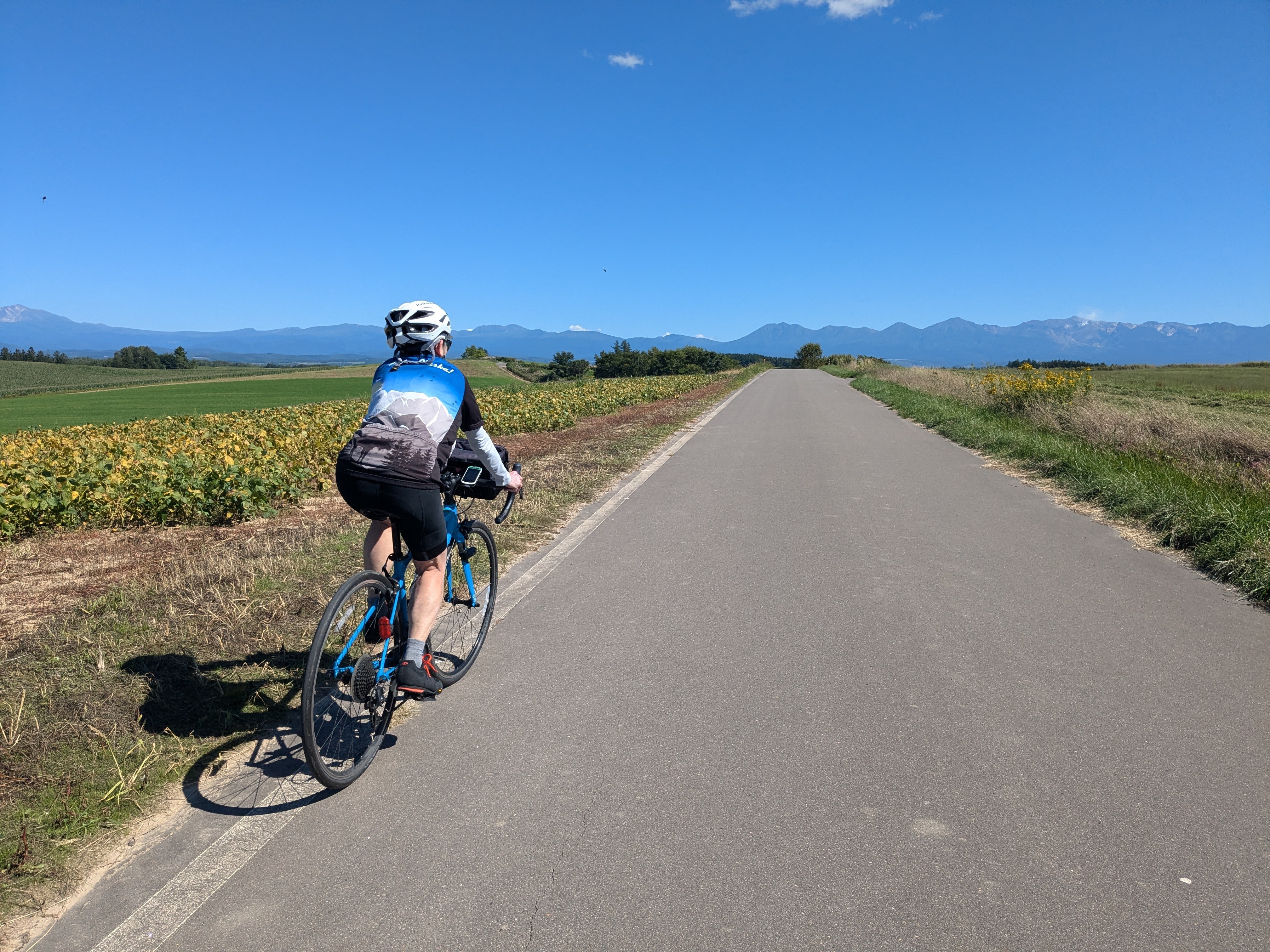 A cyclist rides up to the crest of a hill on a sunny day. The Tokachi Volcanic Group, a mountain range in Hokkaido, stretches across the horizon in the distance.