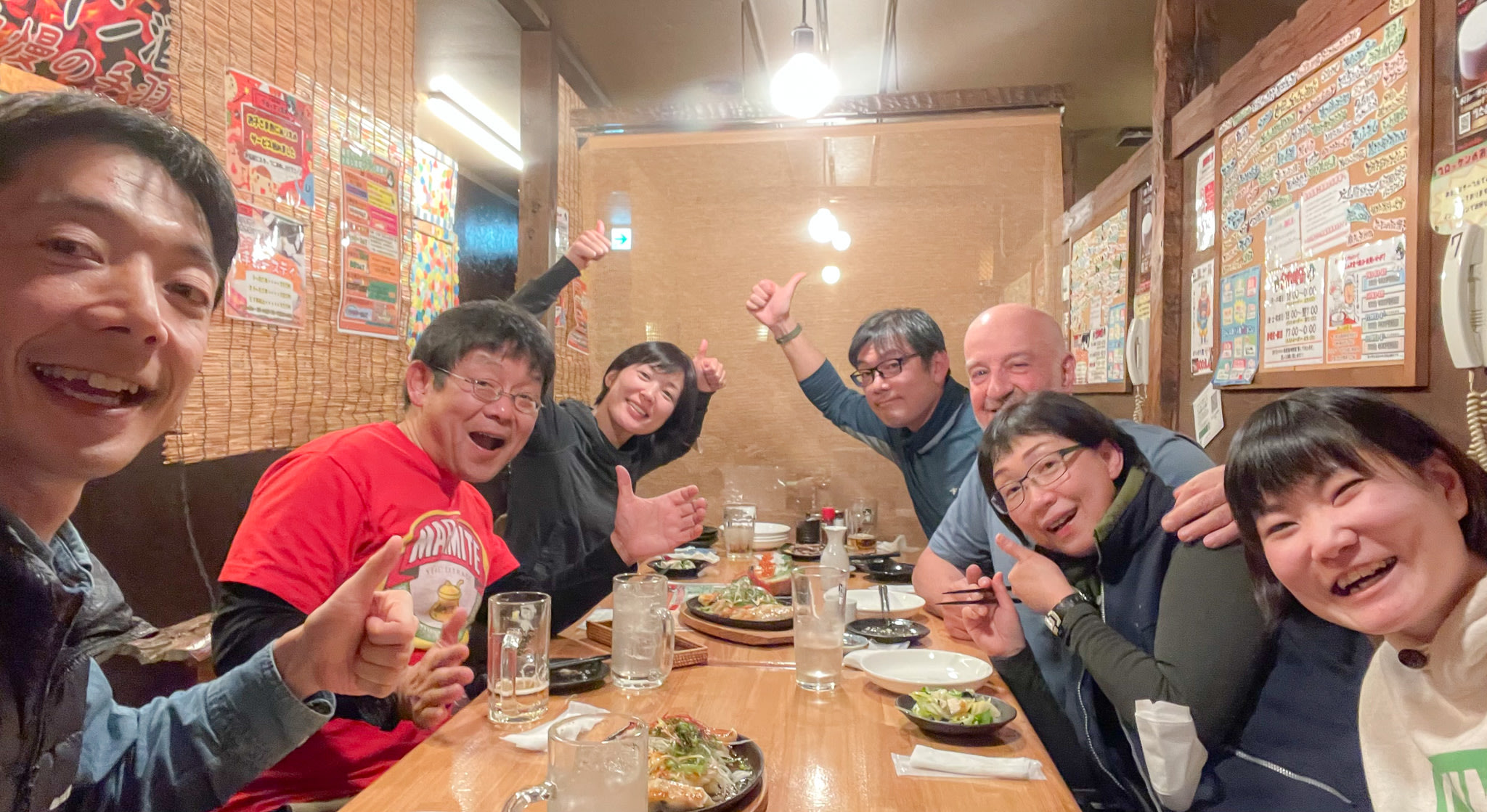 A group of people at a Japanese izakaya enjoying a meal and drinks together. A man at the front of the group is taking a selfie and other members of the group are giving thumbs-ups and waving.
