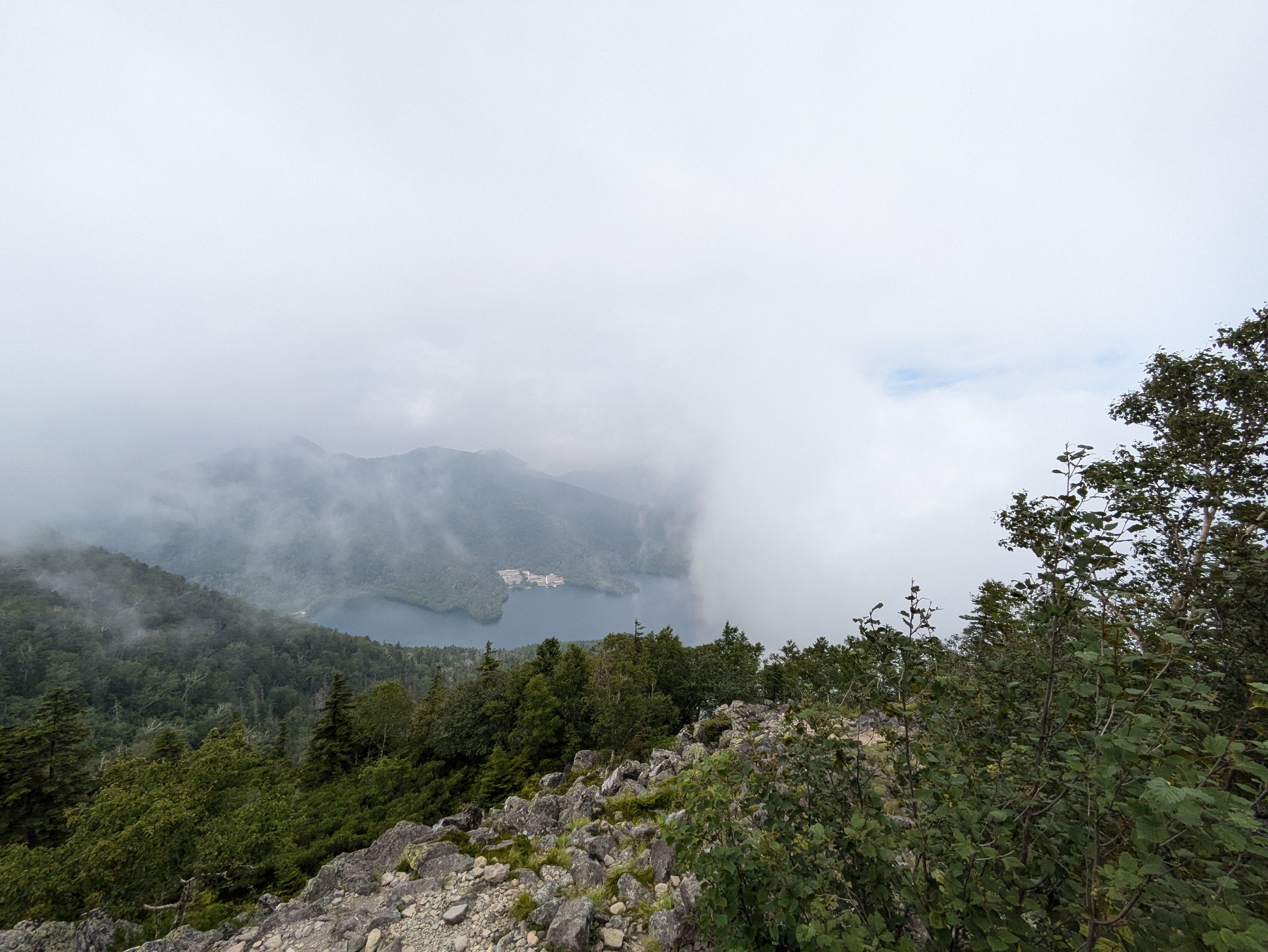 A view of Lake Shikaribetsu from the summit of Mt. Hakuunzan. It is a cloudy day and the view is partially obscured by fog at the summit.