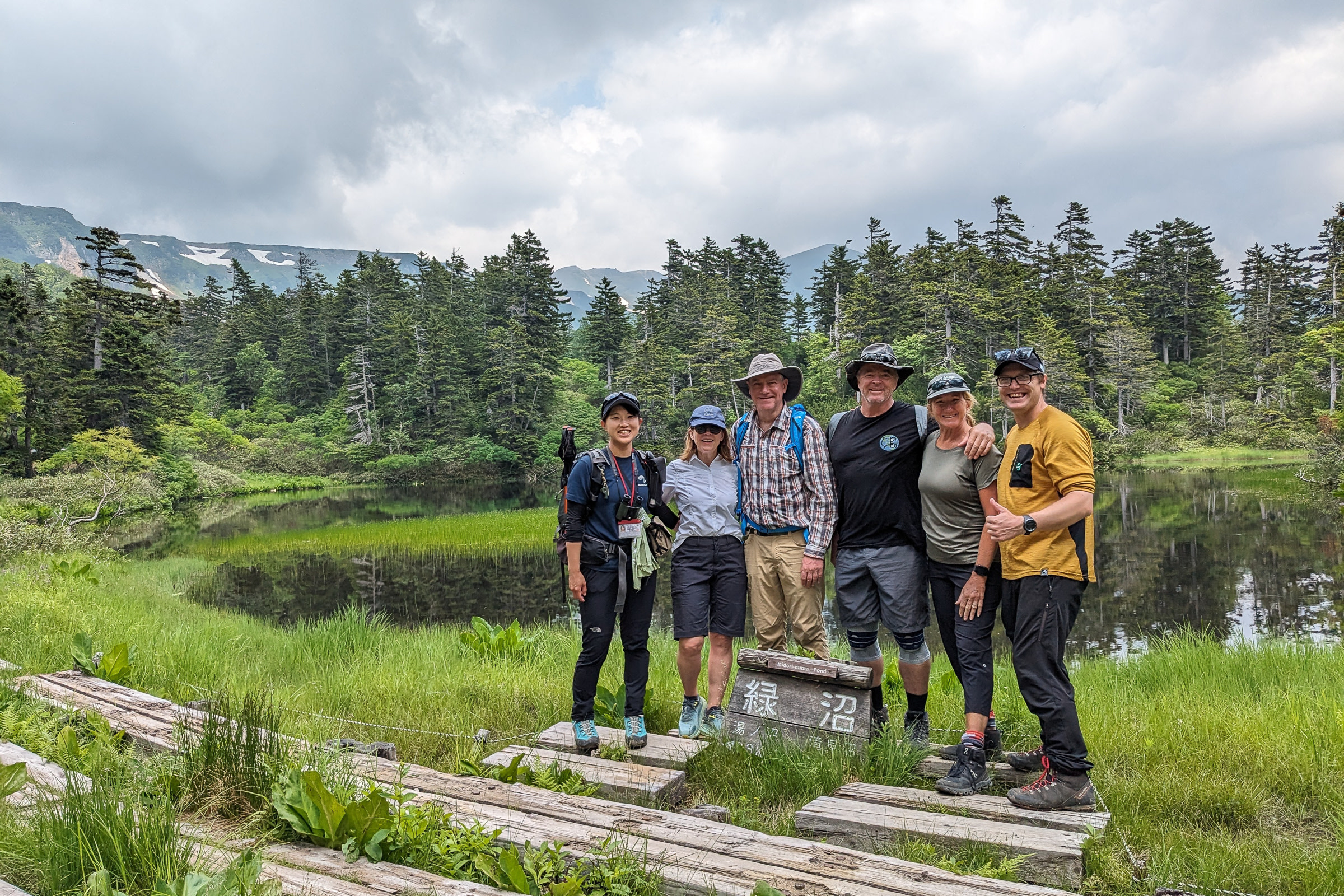 Adventure Hokkaido Guides Yuka and Richard smile with guests. They are standing by a sign reading "Midori Pond" in Japanese with the Midori Pond and alpine scenery behind them.