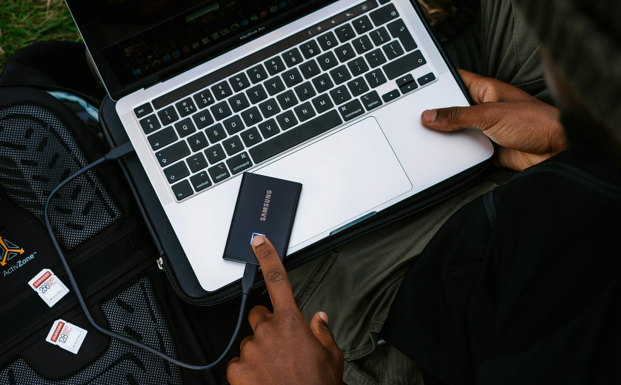 A top-down view of a pair of male hands holding a laptop on the person's lap. They have an SD card reader placed on the laptop next to the track pad. A camera bag and SD storage cards are just at the edge of the image.